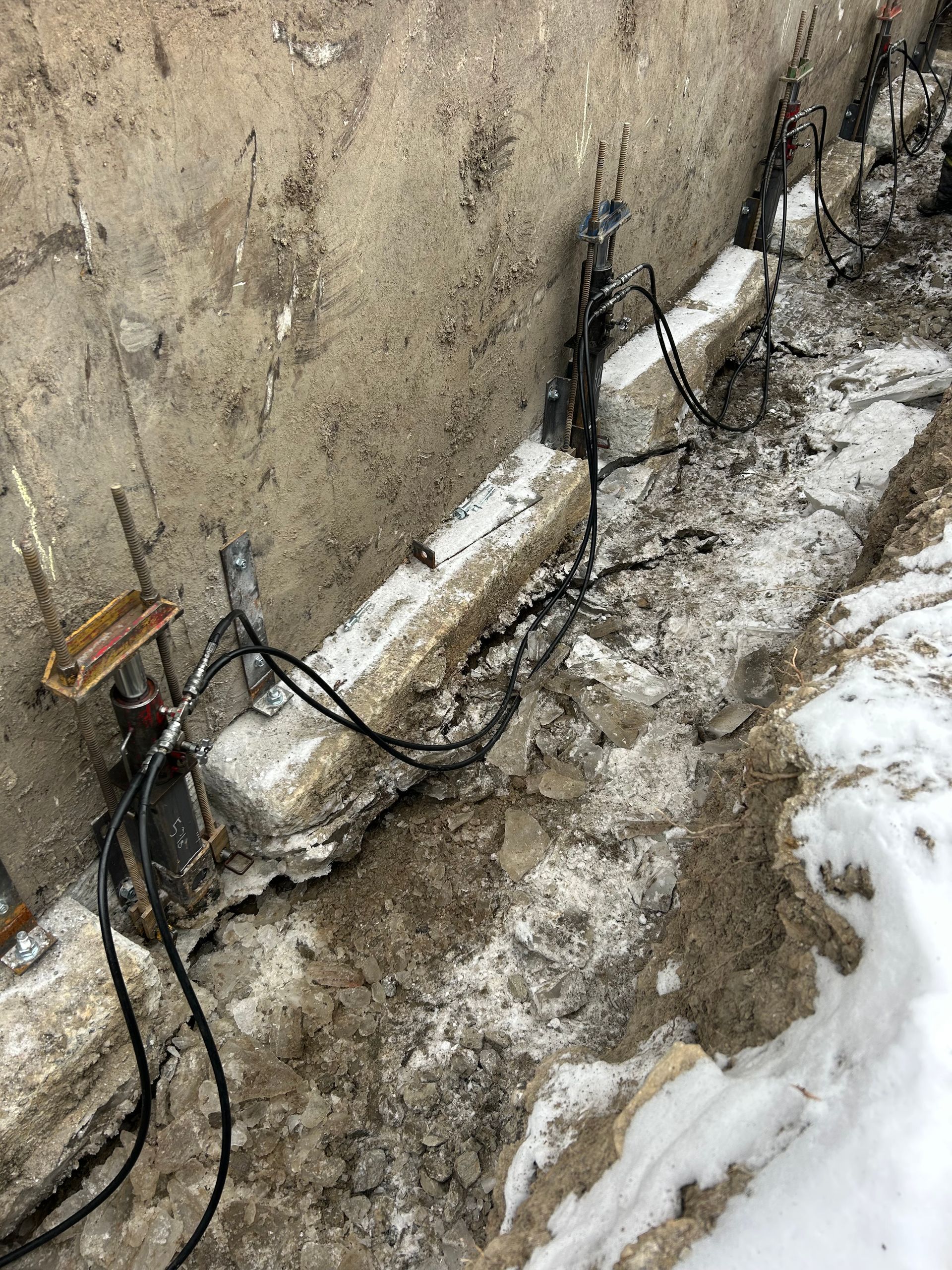 Des vérins hydrauliques sont installés sous un mur de fondation en béton, dans une tranchée boueuse et enneigée, pour assurer le soutien structurel.