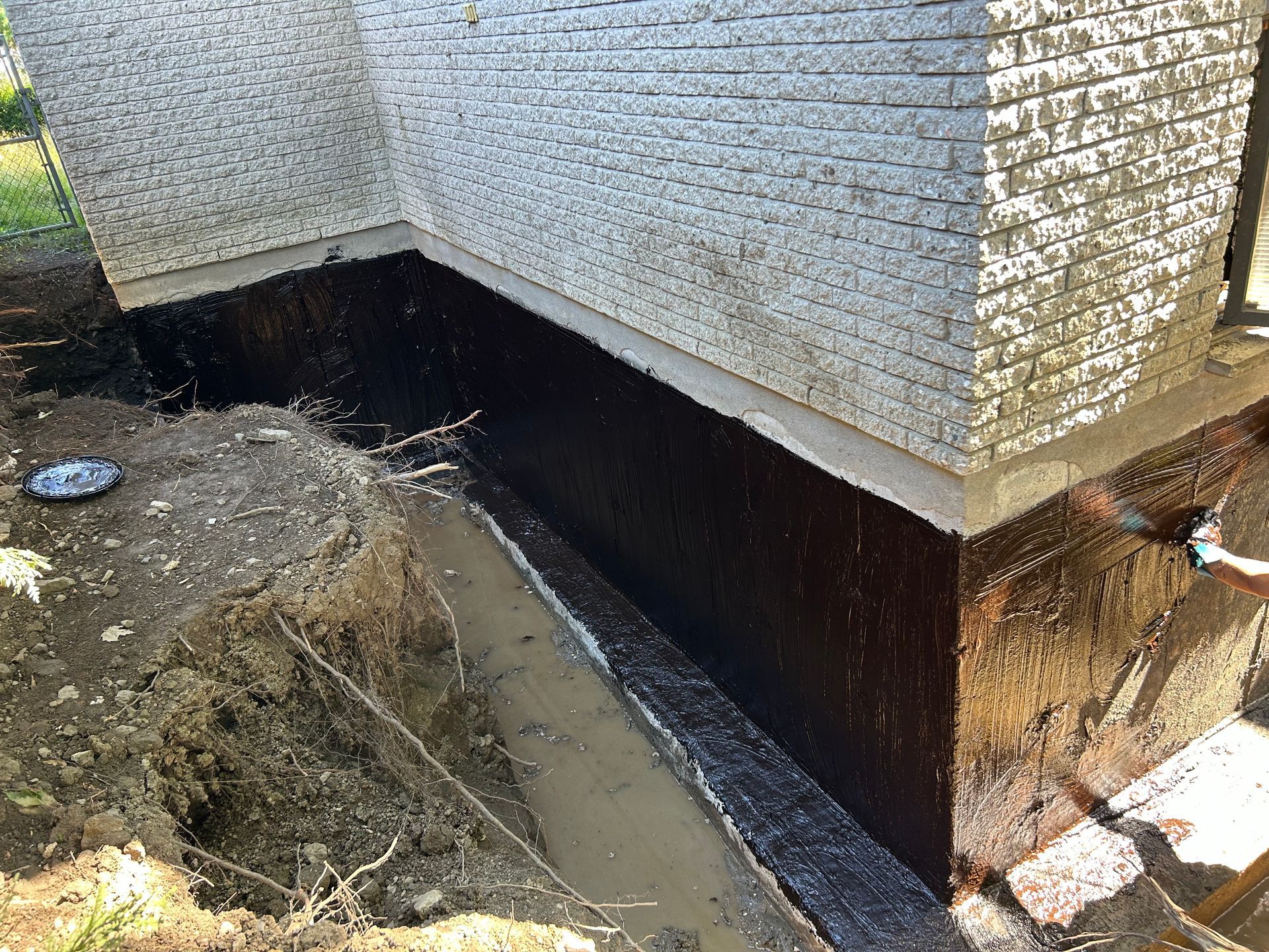 Une tranchée creusée le long des fondations d'une maison en briques blanches, recouvertes d'un enduit imperméable noir.