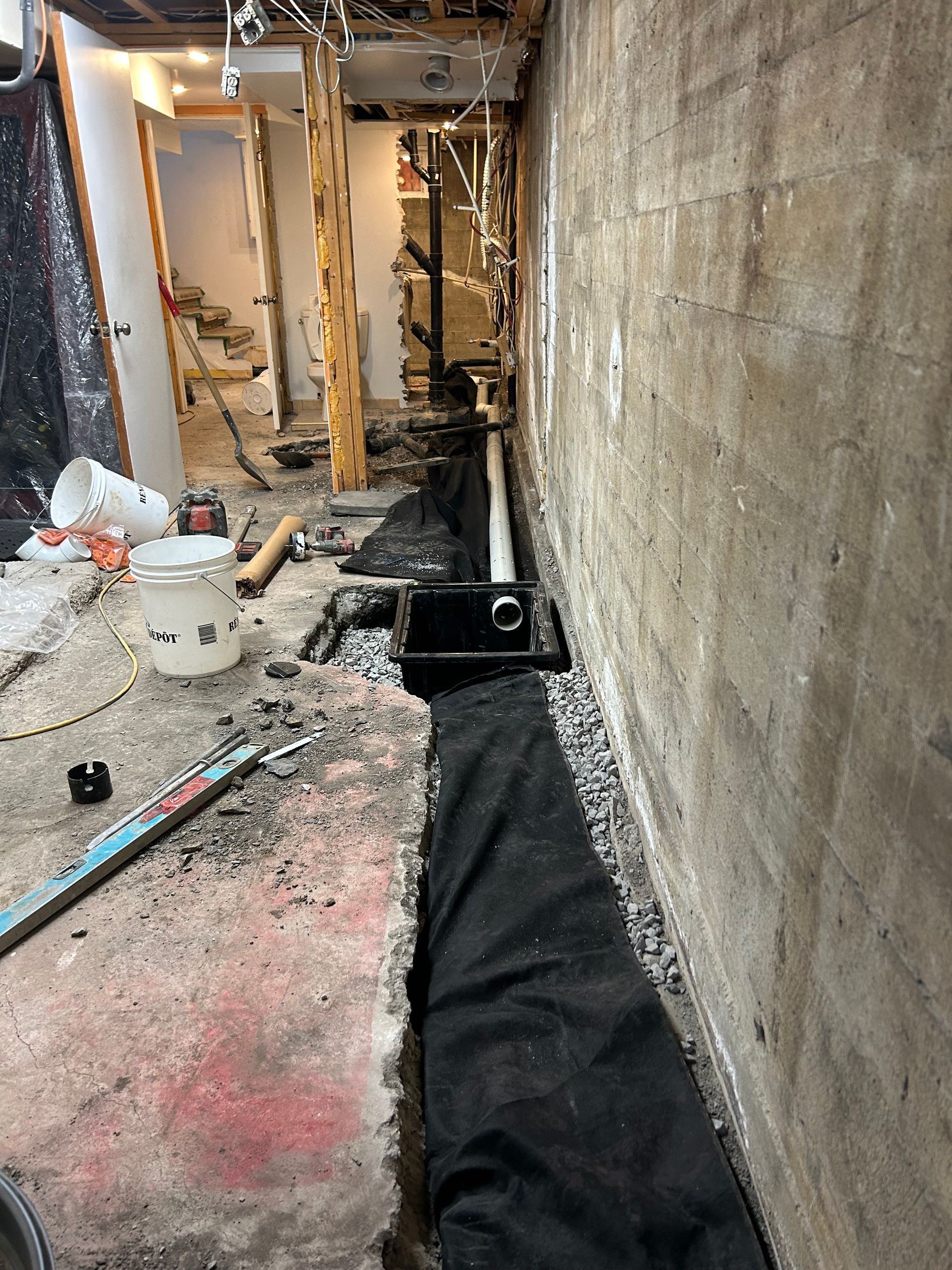 Intérieur d'un sous-sol en rénovation, montrant une tranchée creusée le long d'un mur de fondation en béton avec des tuyaux de drainage et du gravier.