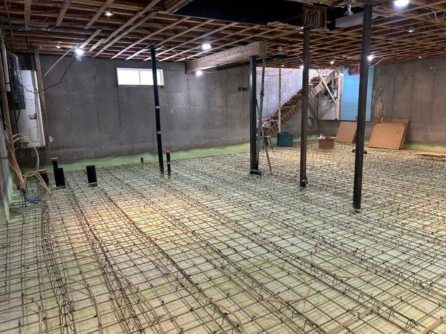 Sous-sol non aménagé avec solives de plafond apparentes, murs en béton et armature métallique préparée pour une nouvelle dalle de béton.