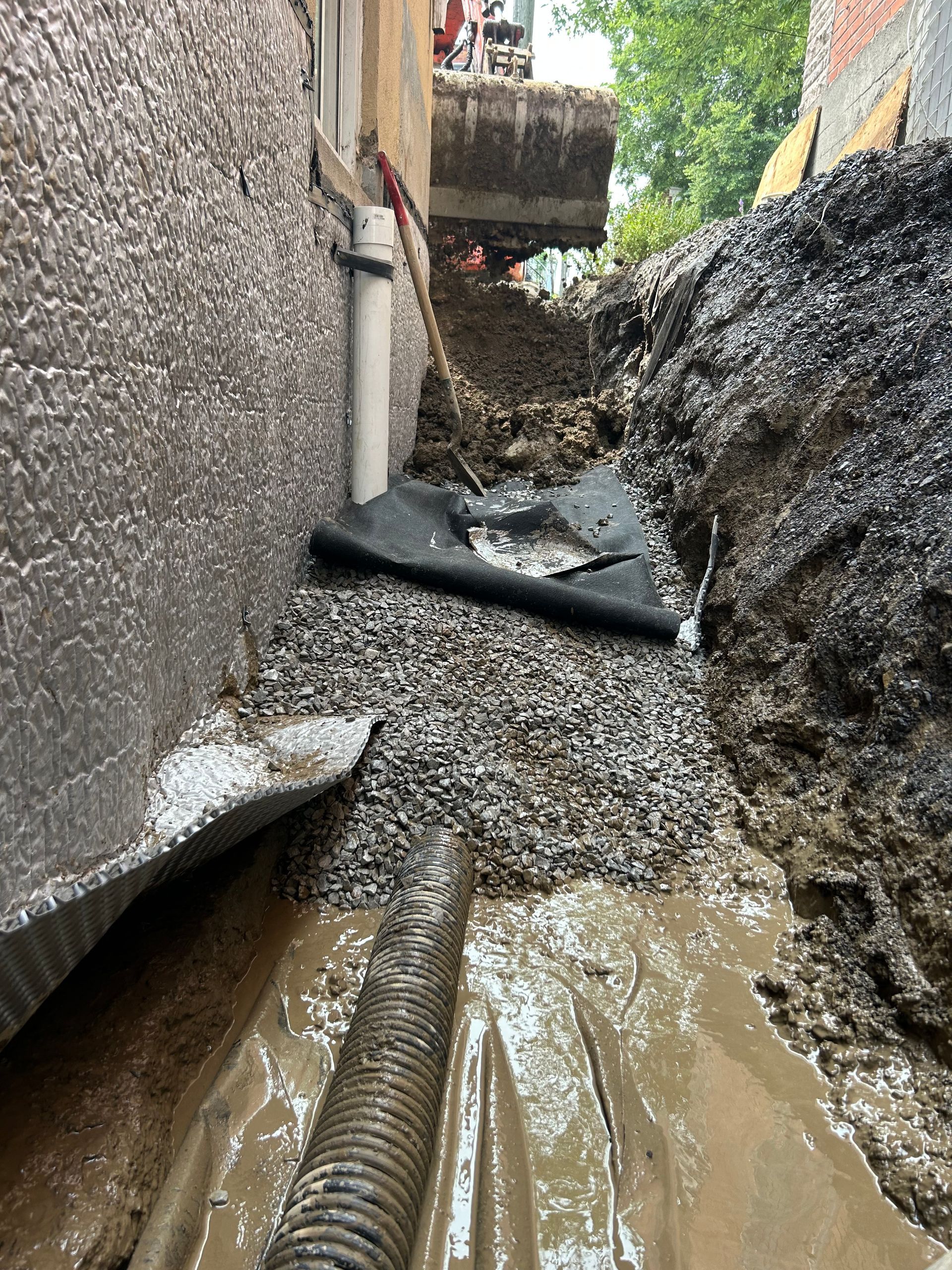 Une tranchée creusée le long des fondations d'un bâtiment, montrant un tuyau de drainage, du gravier et une membrane de protection pour l'étanchéité.