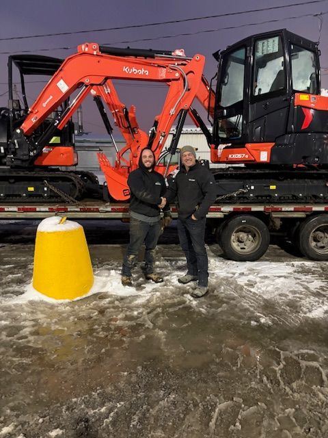 Deux personnes vêtues de vestes sombres se serrent la main devant une excavatrice Kubota orange, posée sur une remorque à plateau, sur un terrain enneigé.