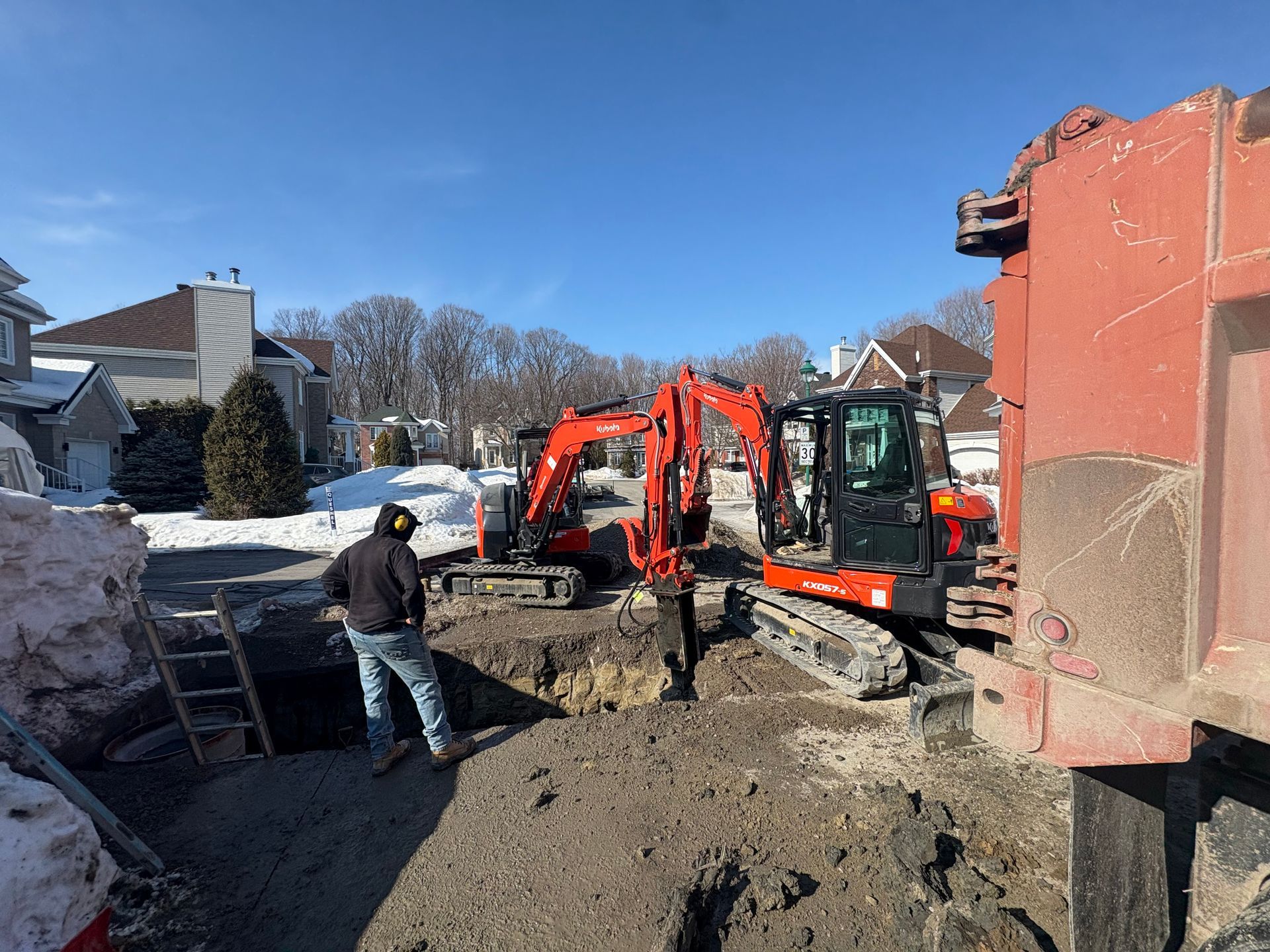 Un ouvrier se tient près d'une tranchée dans un quartier résidentiel où des excavatrices orange effectuent des travaux de construction.