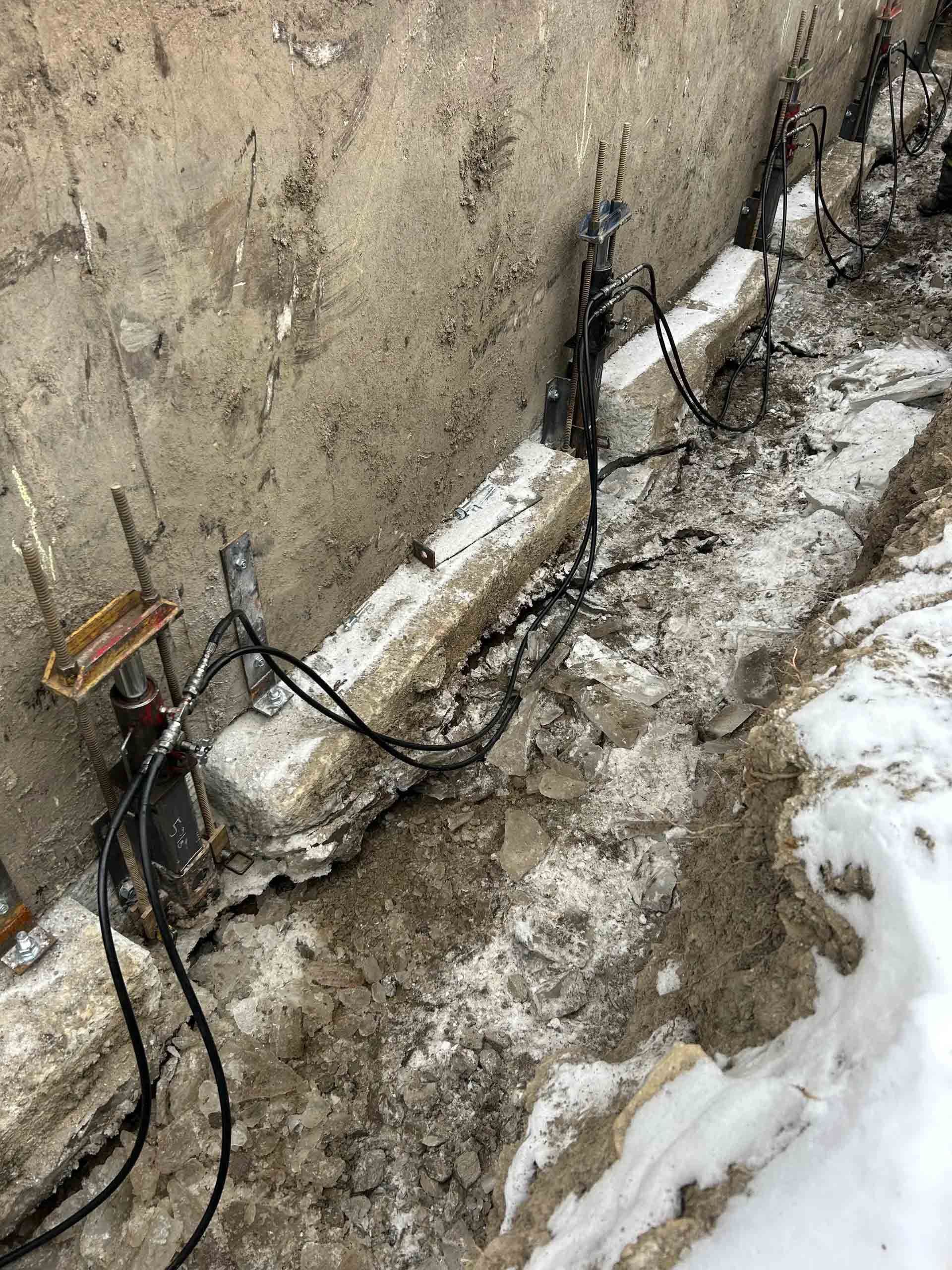 Des vérins hydrauliques sont installés sous un mur de fondation en béton, dans une tranchée boueuse et enneigée, pour assurer le soutien structurel.