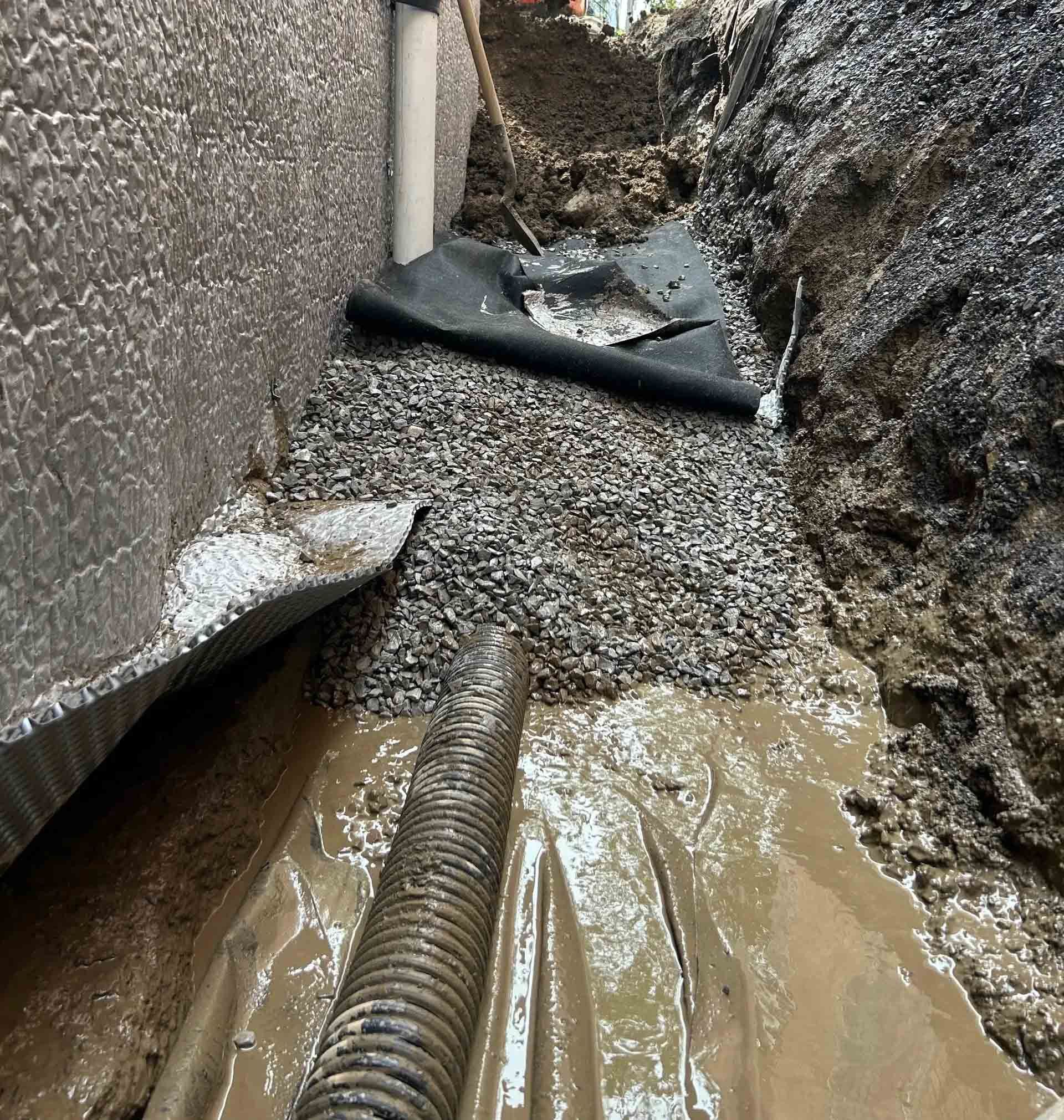 Une tranchée creusée le long des fondations d'une maison, contenant un tuyau de drainage noir, du gravier et une toile de protection pour l'aménagement paysager.