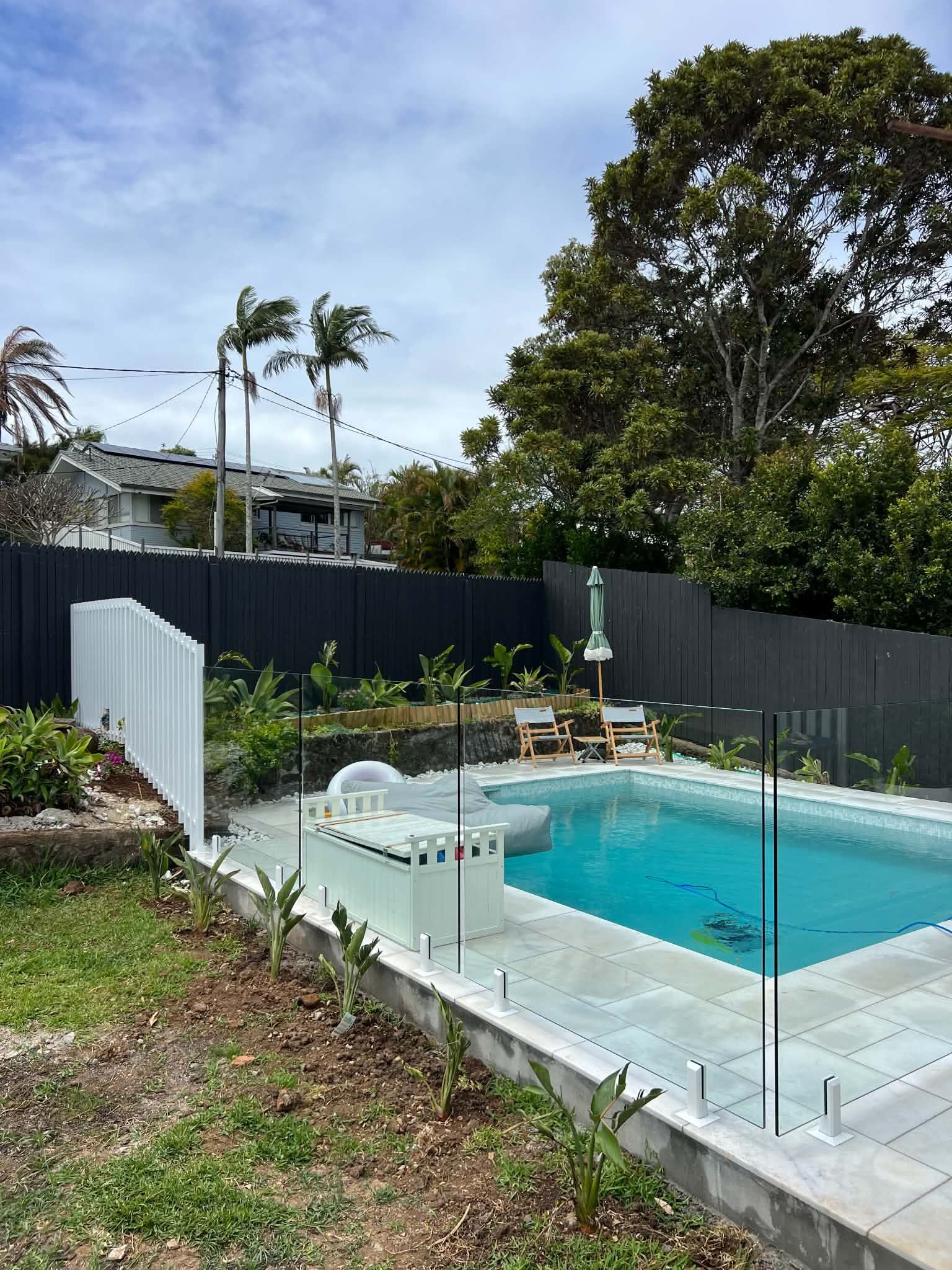Backyard swimming pool with glass fence, lounge chairs, and trees under a clear sky