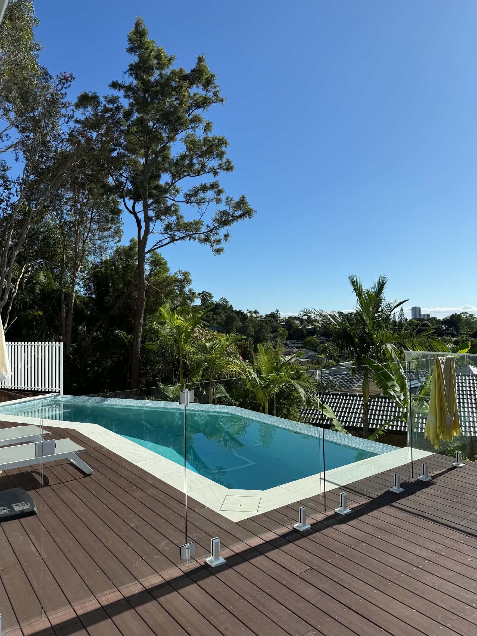 Backyard pool with wooden deck, glass railing, and trees under a clear blue sky