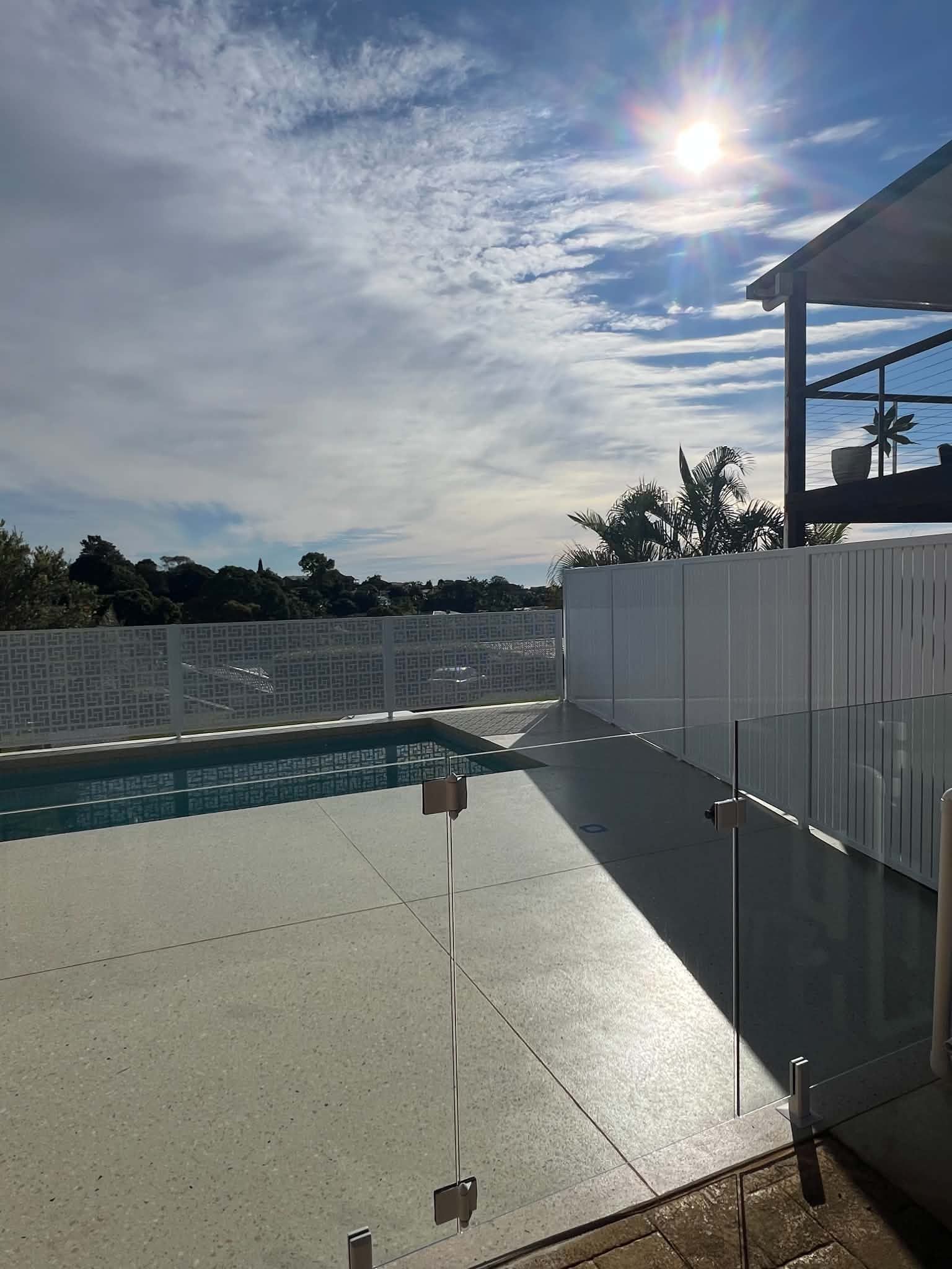 Sunny poolside patio with white fence, reflecting water, and a cloudy sky