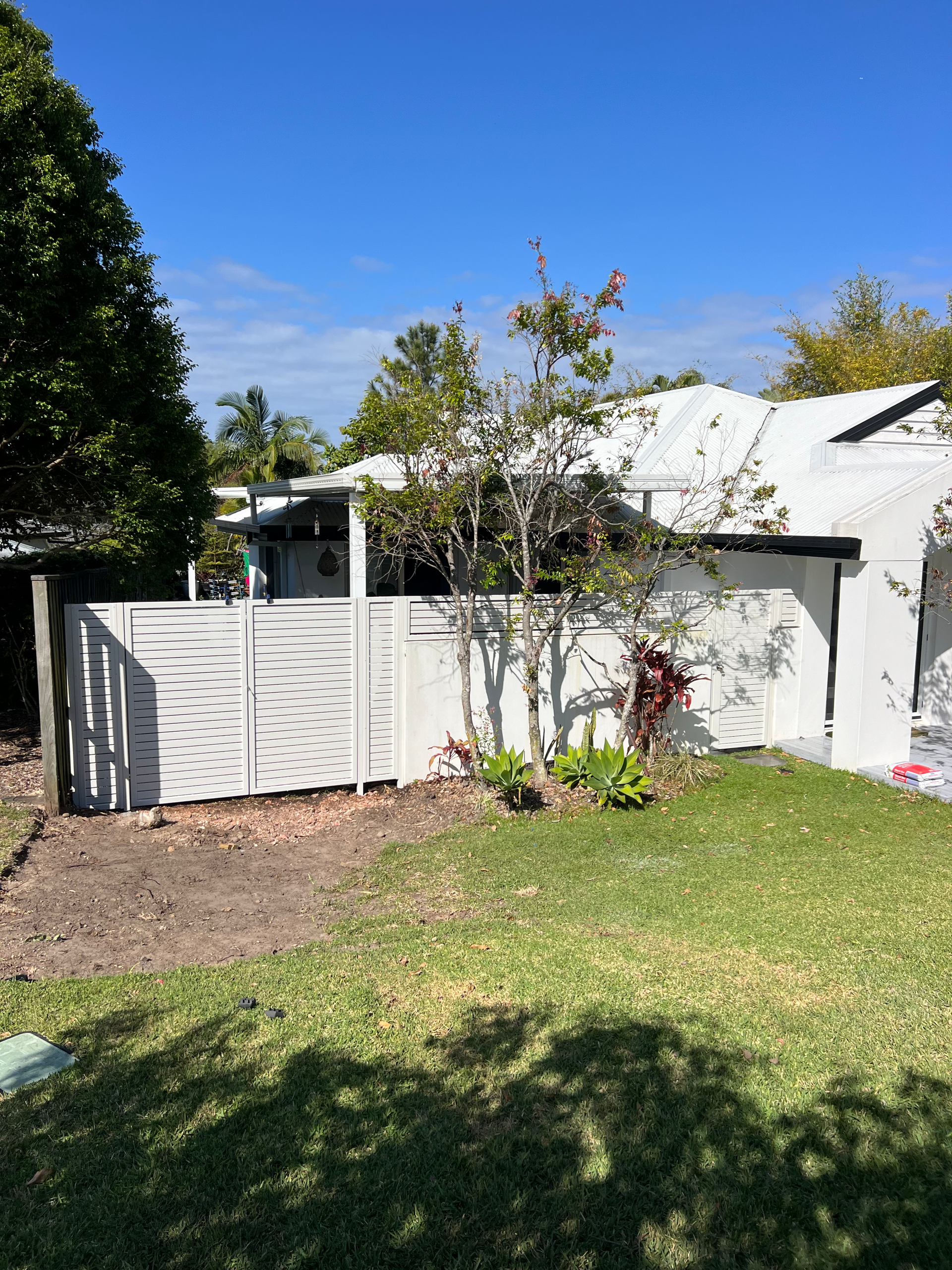 Backyard with white fence, small tree, and green lawn under a bright blue sky