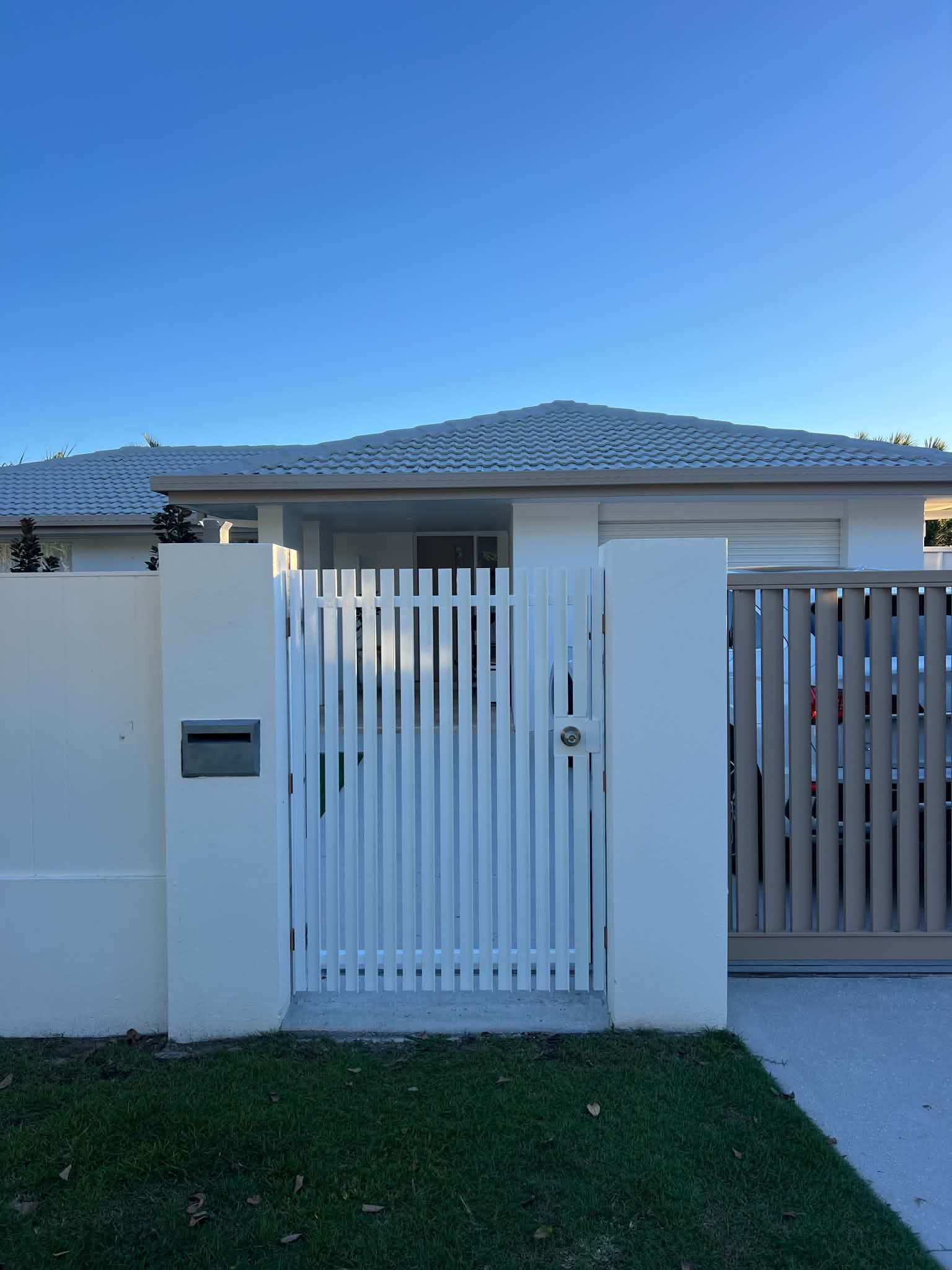 White front gate with vertical slats and adjacent wall in front of a single-story house under a blue sky