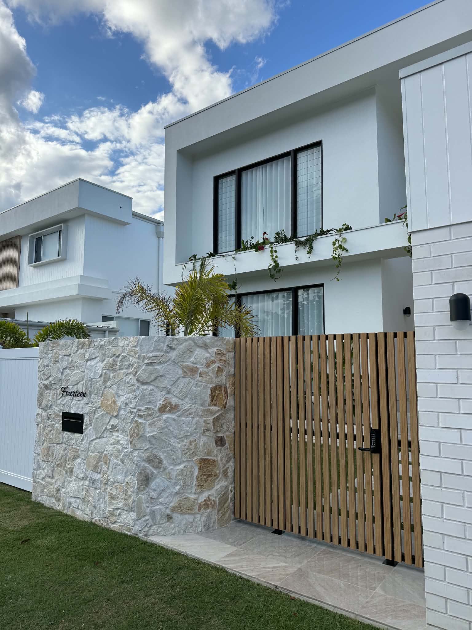 Modern white two-story house with stone wall, wooden gate, and small front lawn under a blue sky
