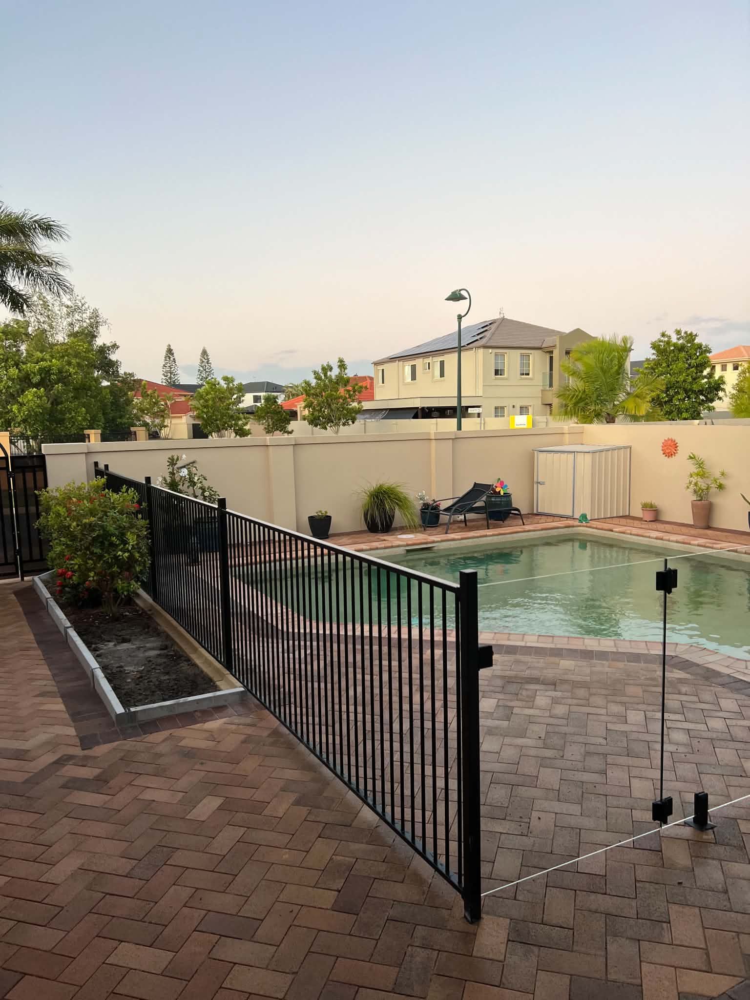 Backyard pool area with black fence, brick patio, and beige house at dusk
