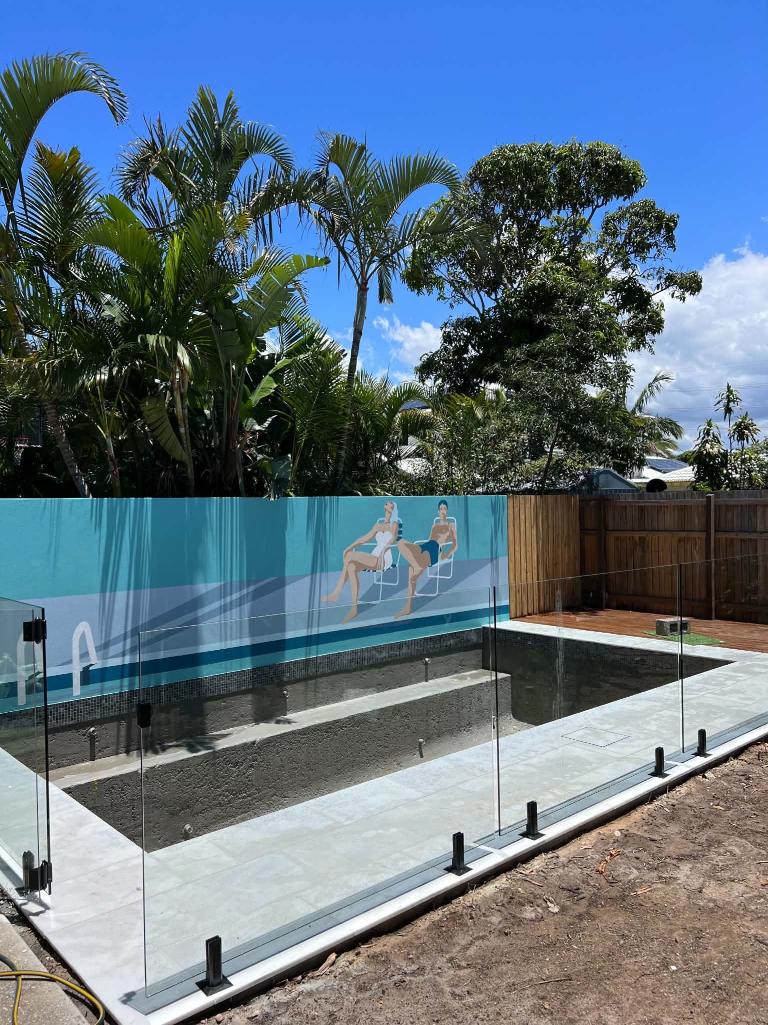 Backyard pool under construction with glass fencing, mural wall, and palm trees behind it