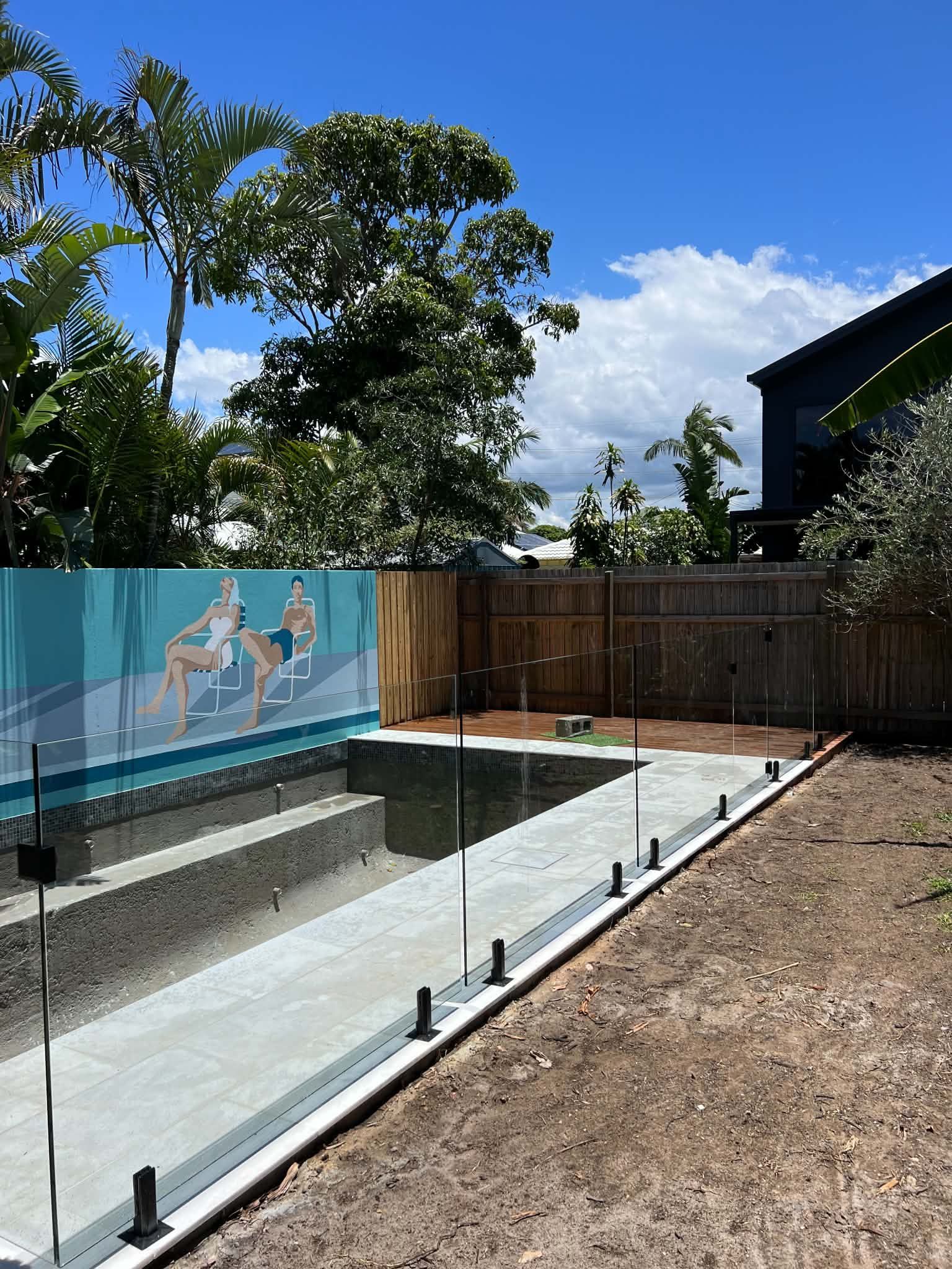 Backyard pool under construction with concrete deck, glass fence, mural wall, and trees beyond