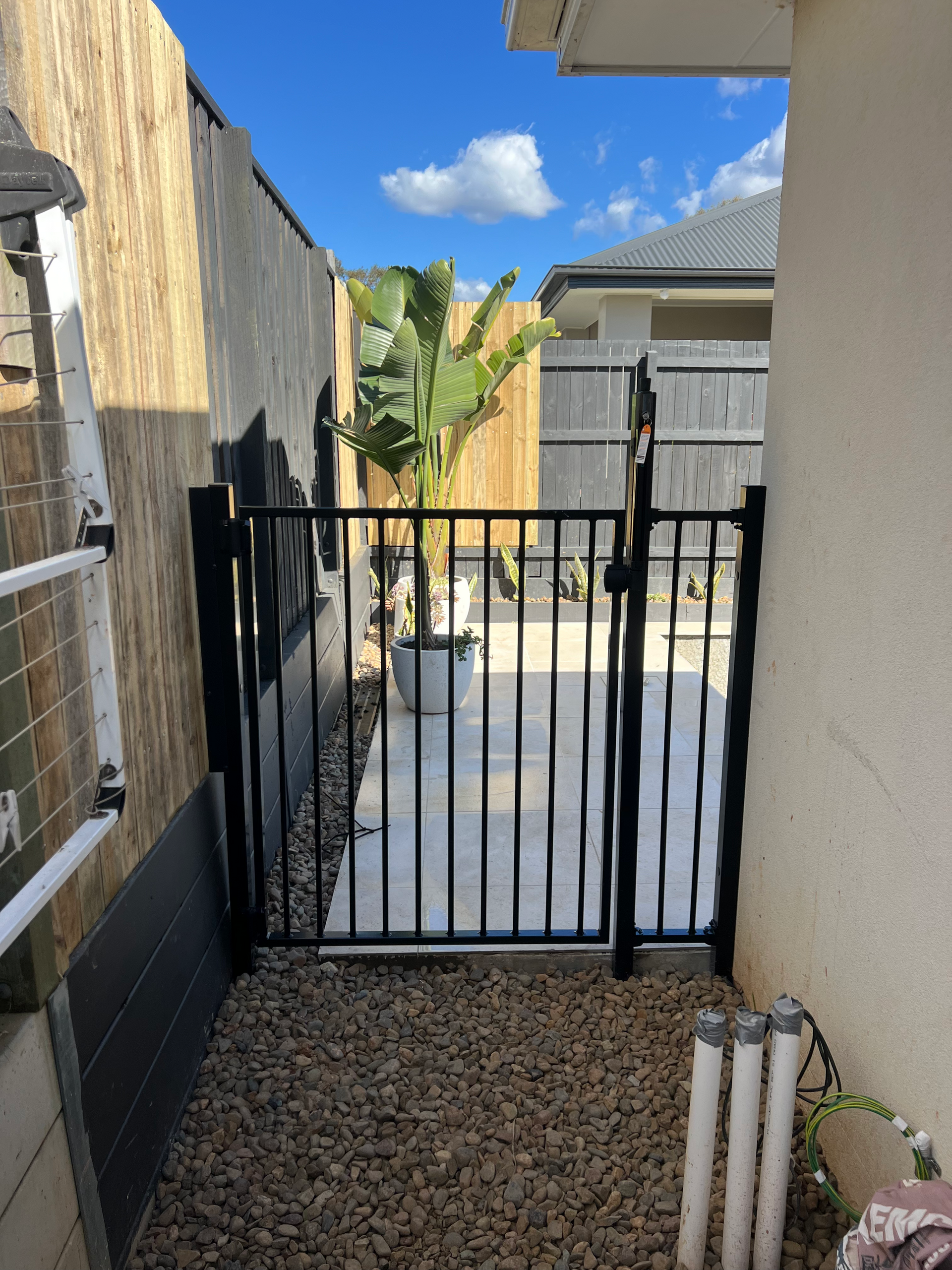Narrow side yard with black metal gate, gravel path, wooden fence, and potted plants leading to a backyard.