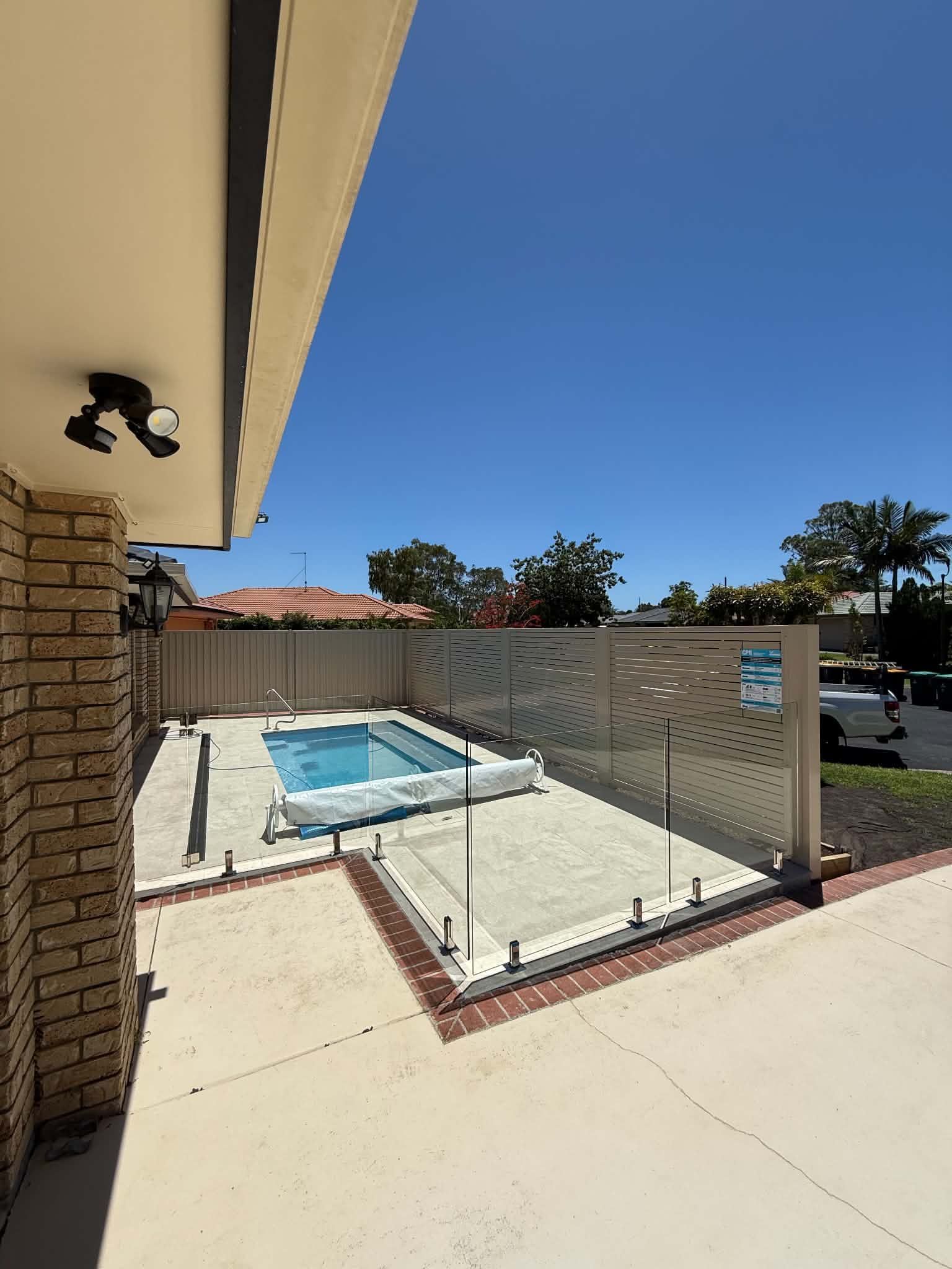 Backyard pool with a white patio, chain-link fence, and house roof overhang in bright sun