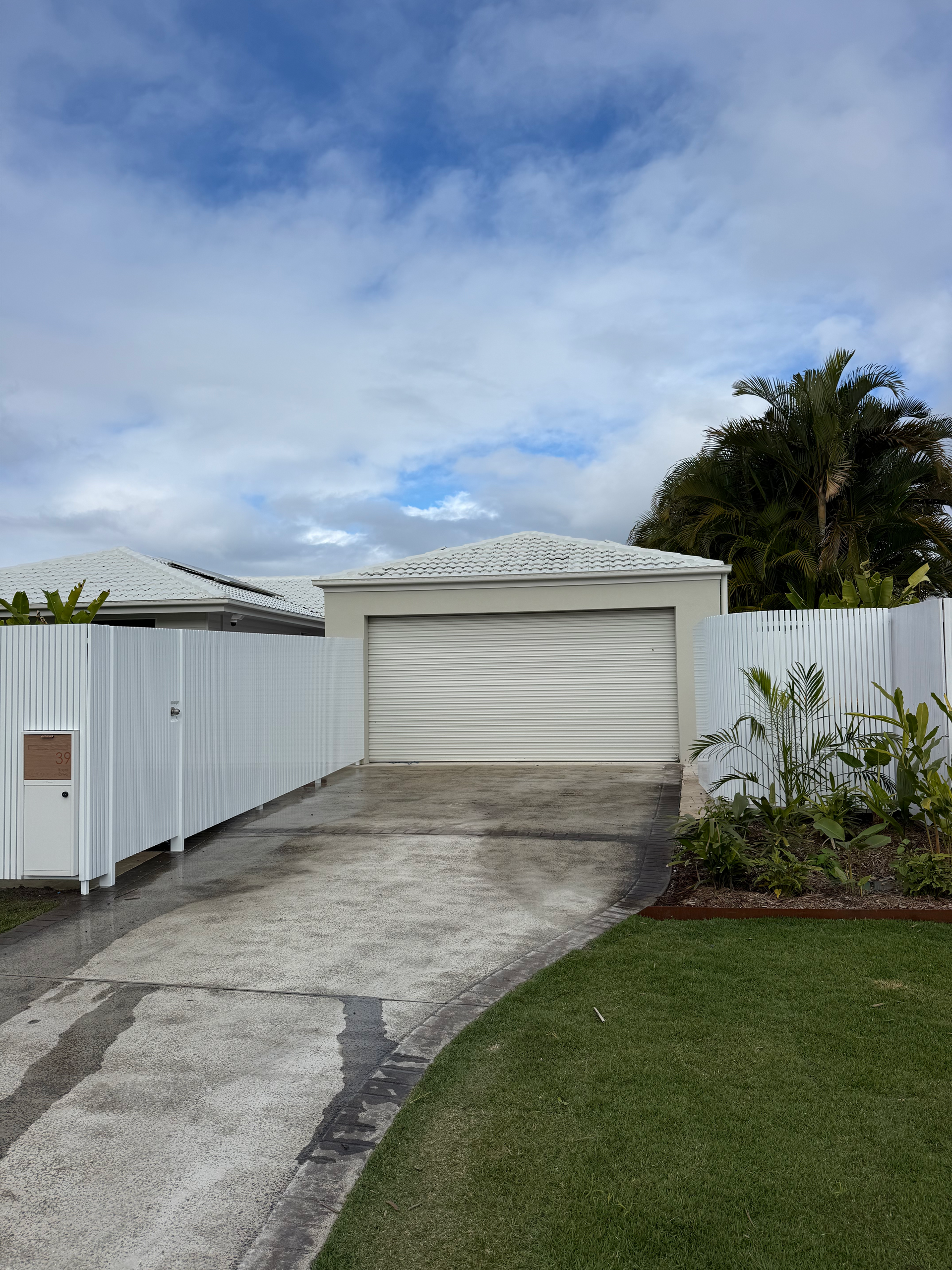 Driveway leading to a white garage beside a white fence and lawn under a cloudy sky.