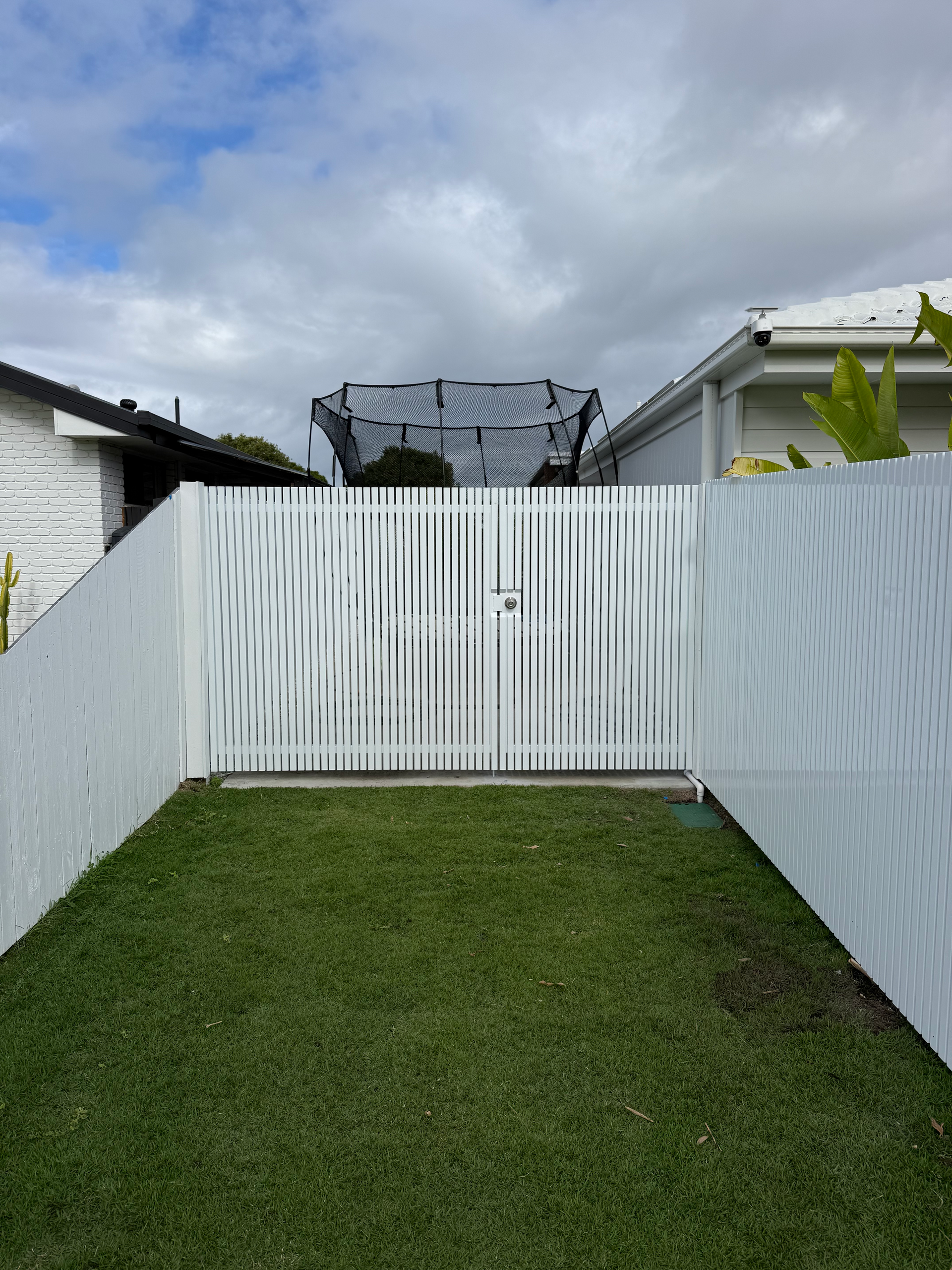Narrow grassy backyard with white fences and gate under a cloudy sky