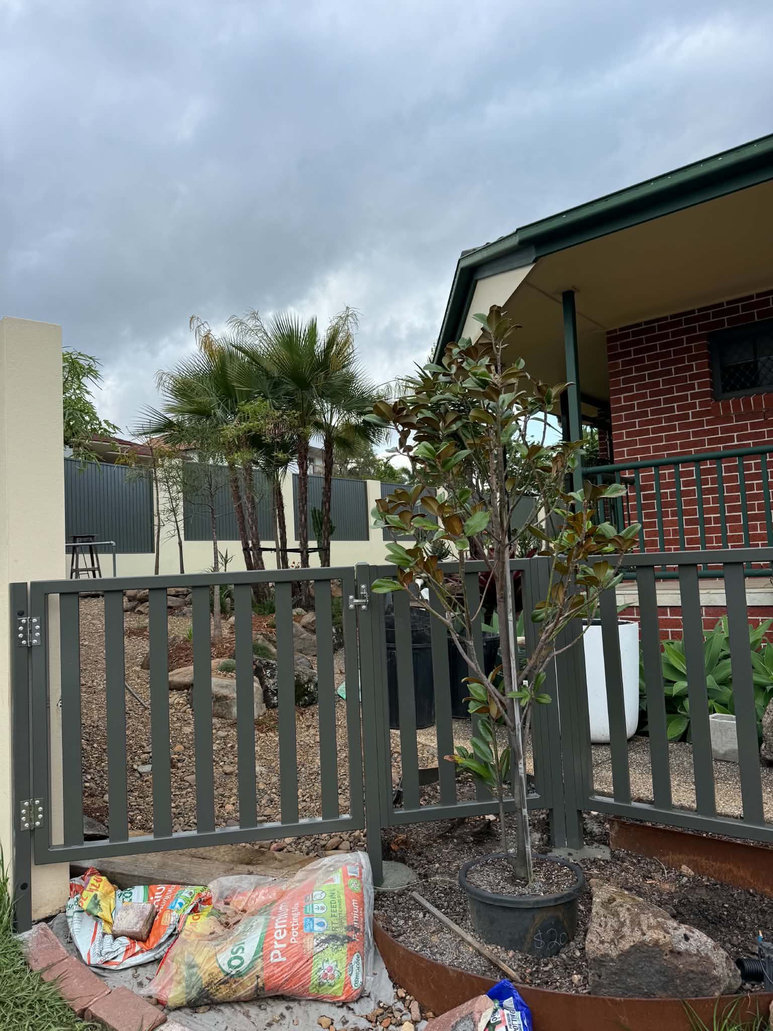 Front yard with gray fence, small trees, and a colorful bag of garden debris near a brick house