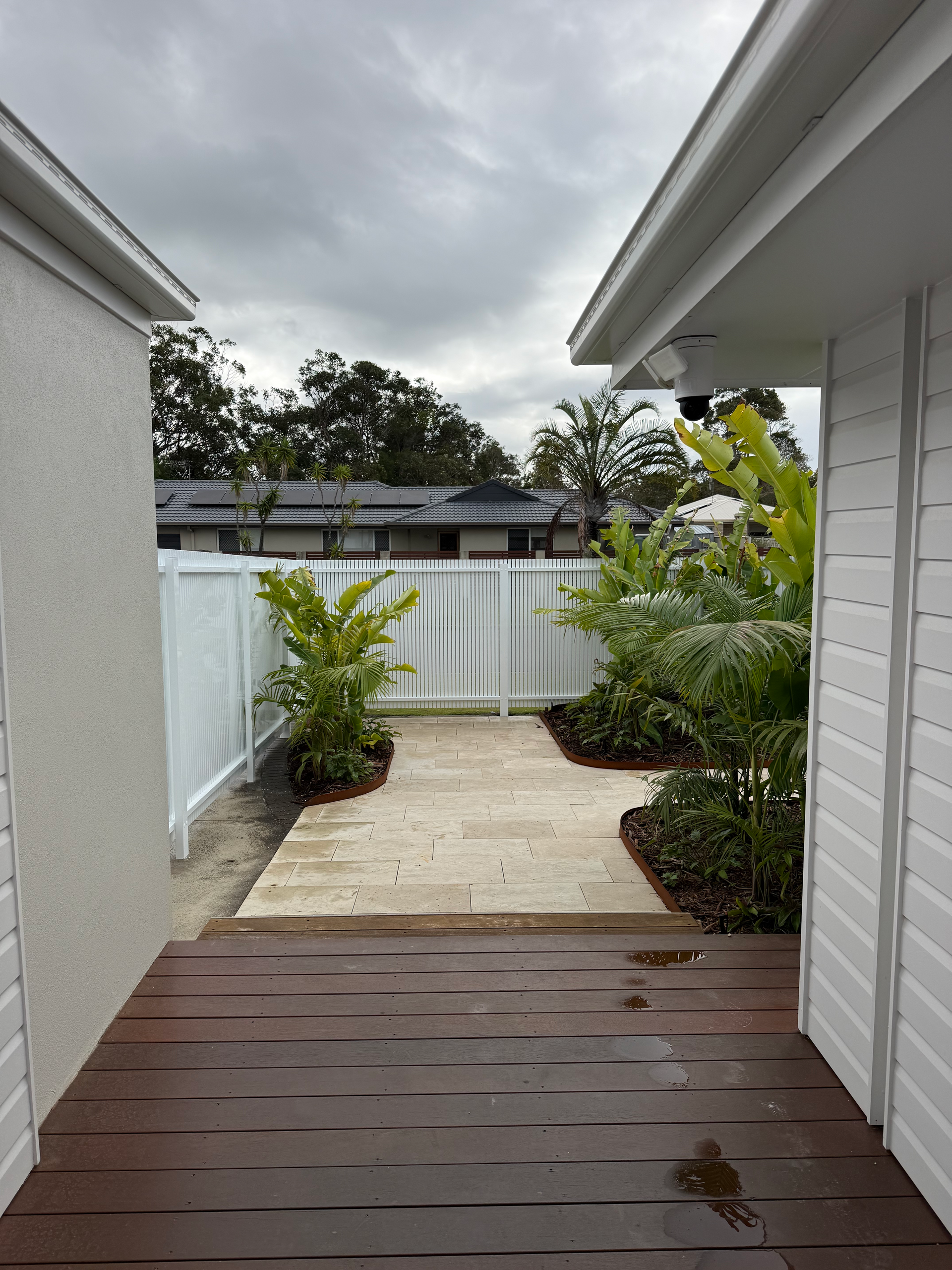 Narrow backyard walkway between white walls, with pavers, plants, and a cloudy sky beyond.