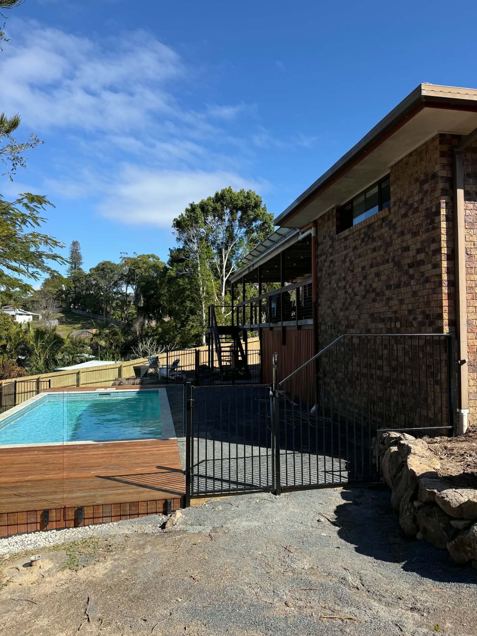 Backyard pool beside a brick house, with a wooden deck, black gate, and blue sky.