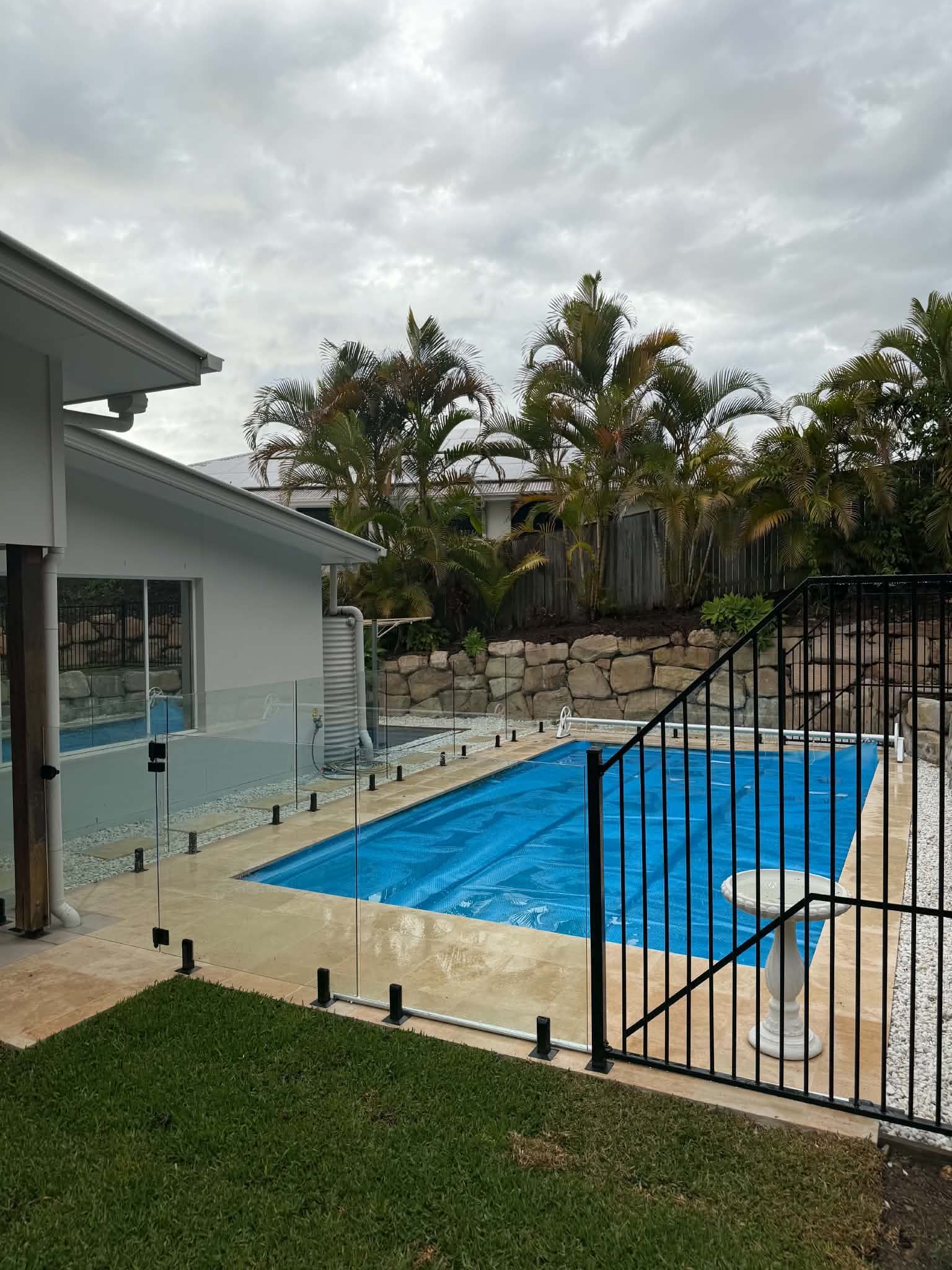 Backyard pool with blue cover beside a house and fenced patio under cloudy skies