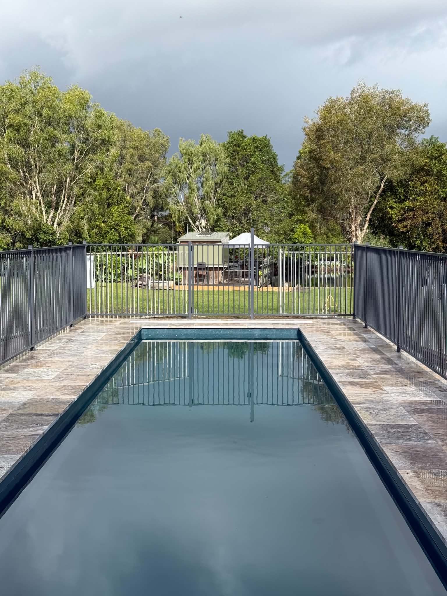 Outdoor swimming pool with calm water, stone deck, and trees under a cloudy sky