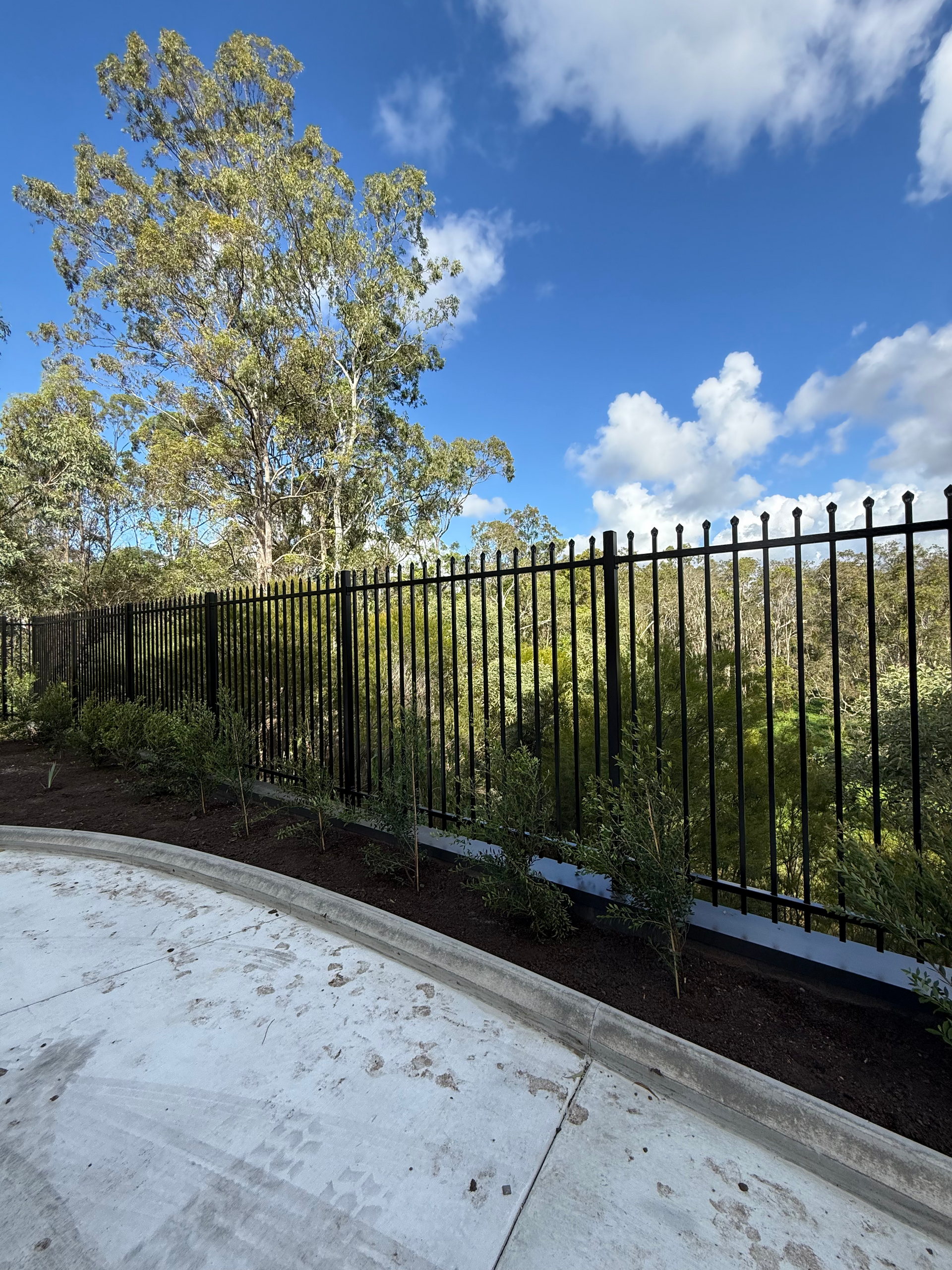 Black metal fence beside a curved concrete path, with trees, greenery, and a blue sky with clouds.