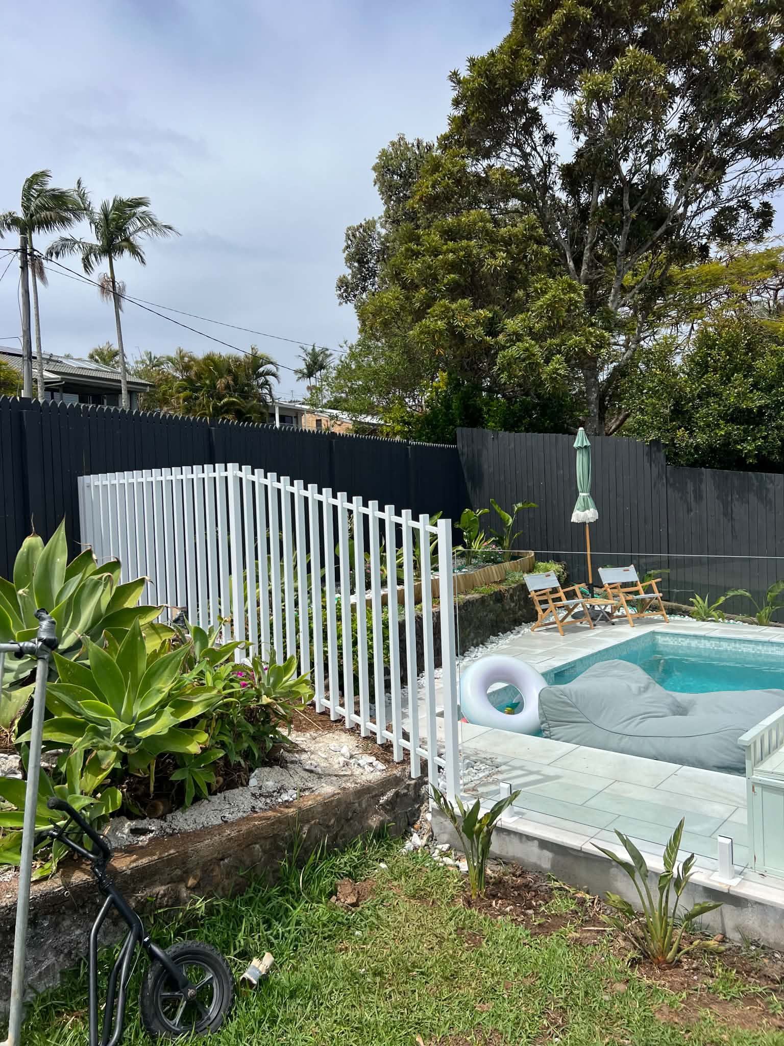 Backyard pool beside white fence and tropical plants on a sunny day