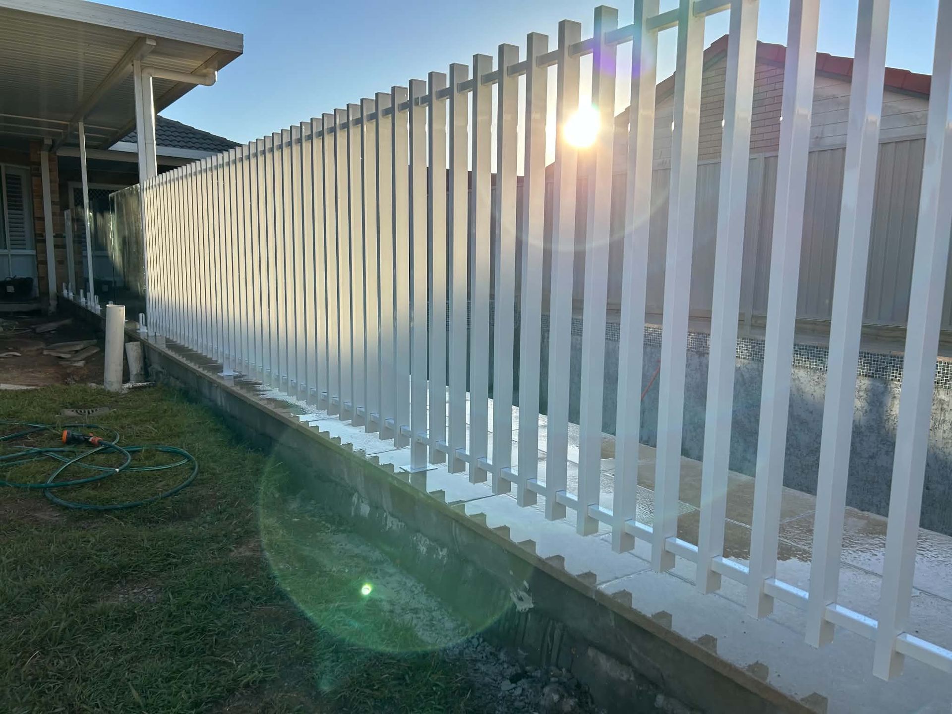 White metal fence along a yard at sunset, with sun glare and a house on the left