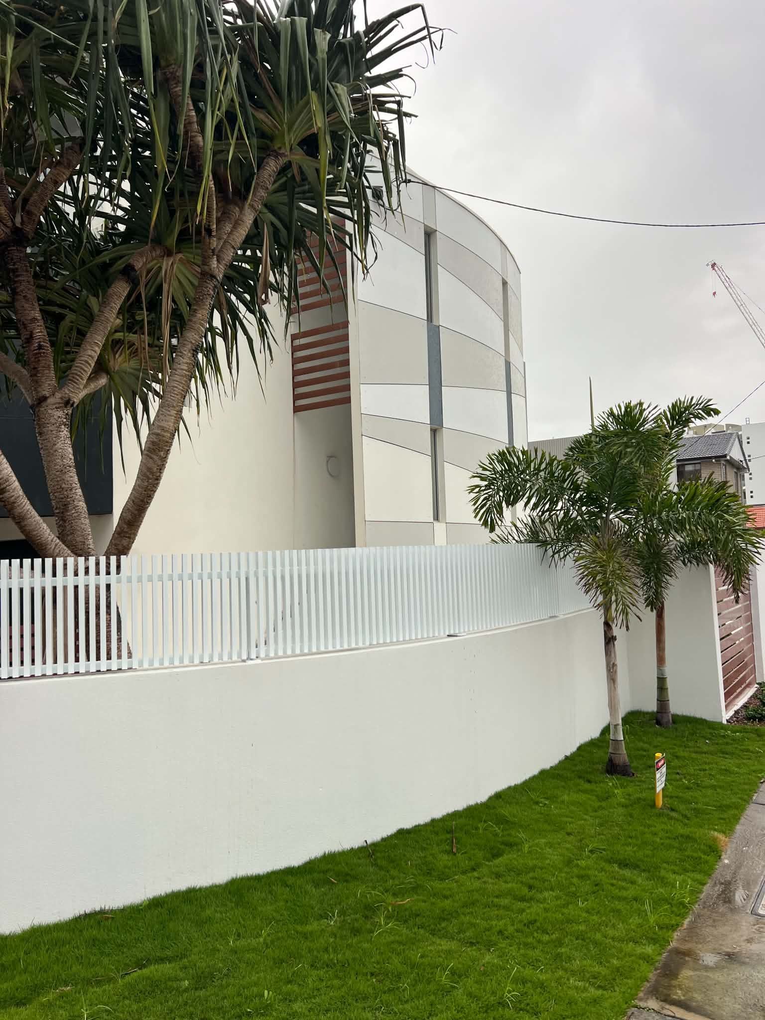 White curved wall with picket fence, tall trees, and green grass beside a sidewalk under a cloudy sky