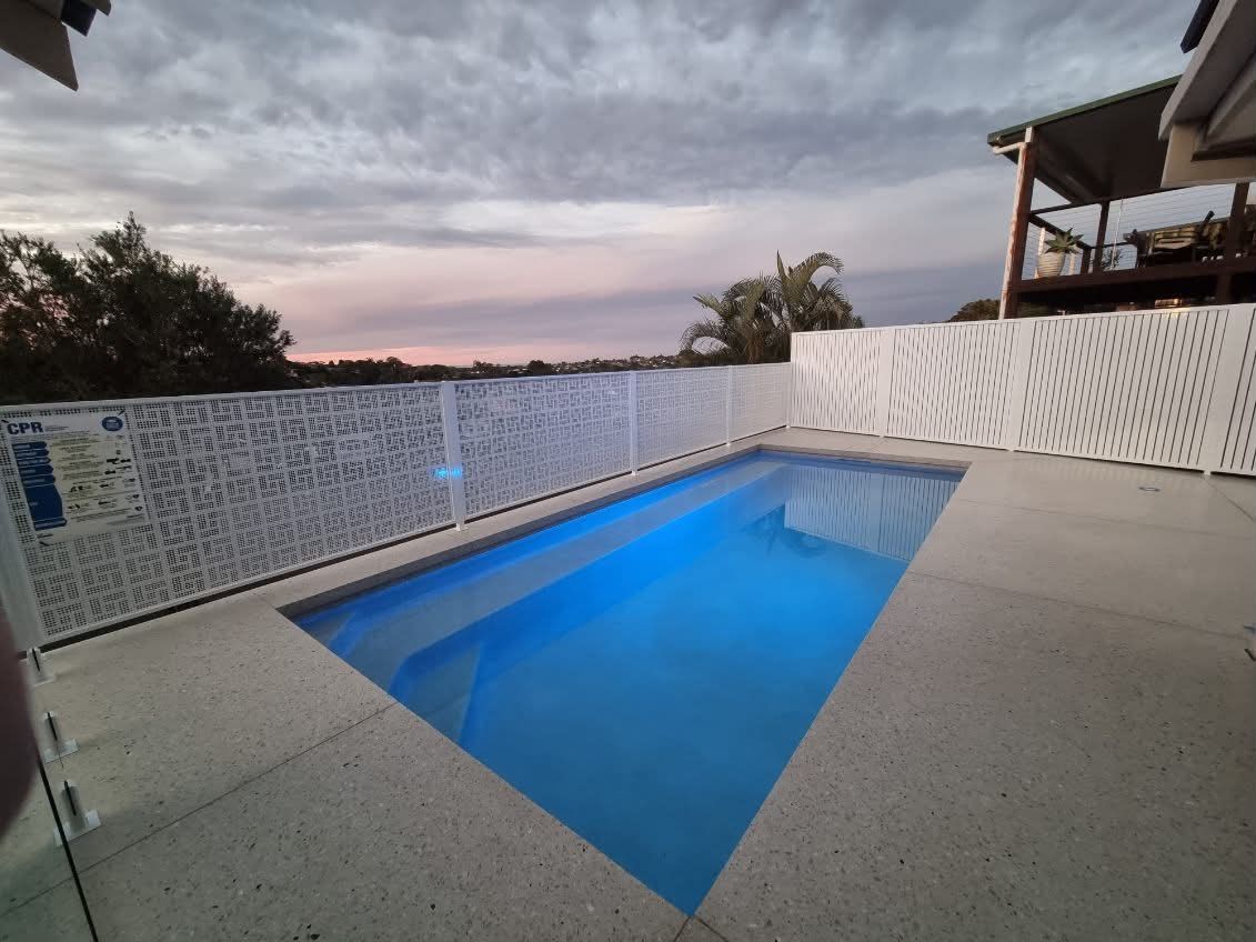 Backyard swimming pool at dusk with blue water and patterned white fence panels