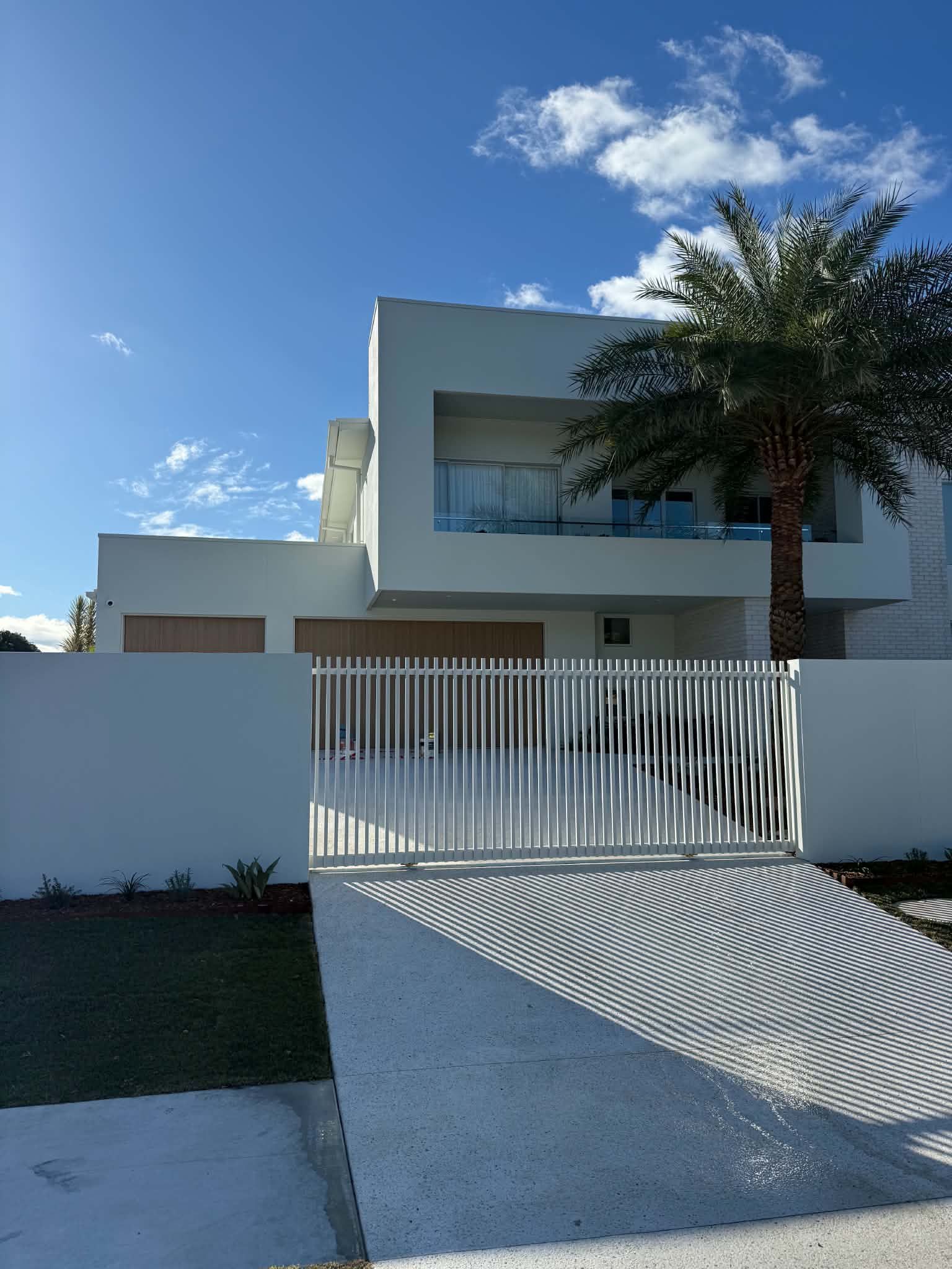 Modern white two-story house with glass balcony, palm tree, and gated driveway under a blue sky