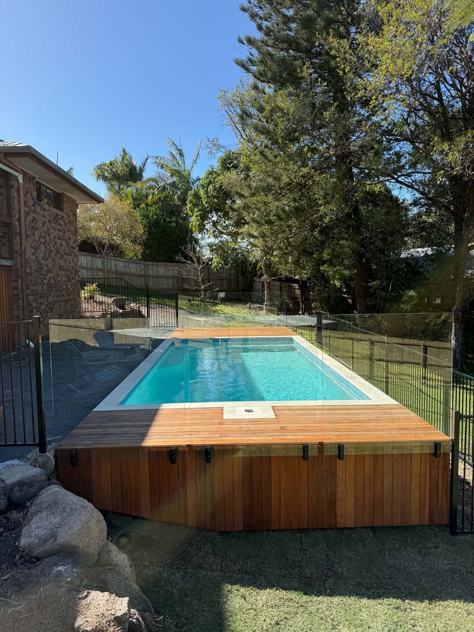 Backyard pool with wooden deck, black safety fence, and trees under a clear blue sky