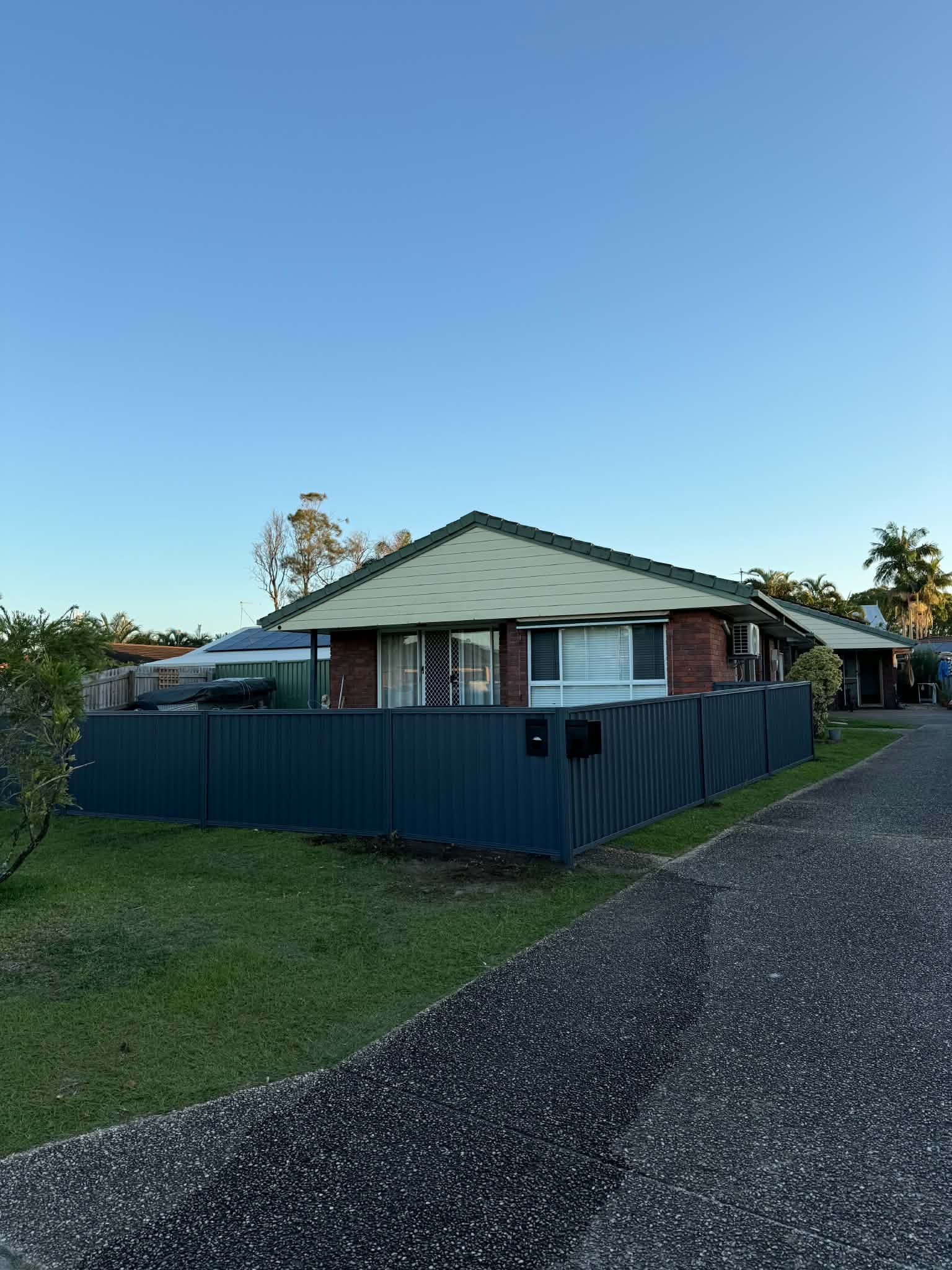 Single-story house with a green roof and blue fence beside a gravel driveway at dusk