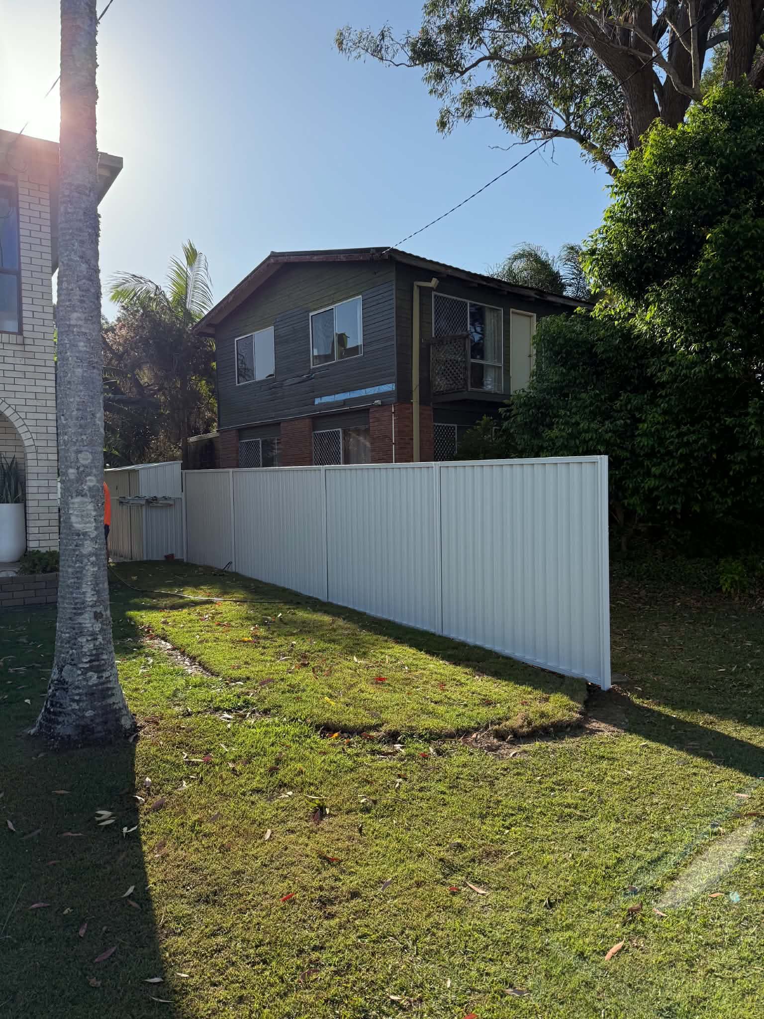 Backyard with white fence, two-story house, trees, and sunlight casting shadows on the grass.