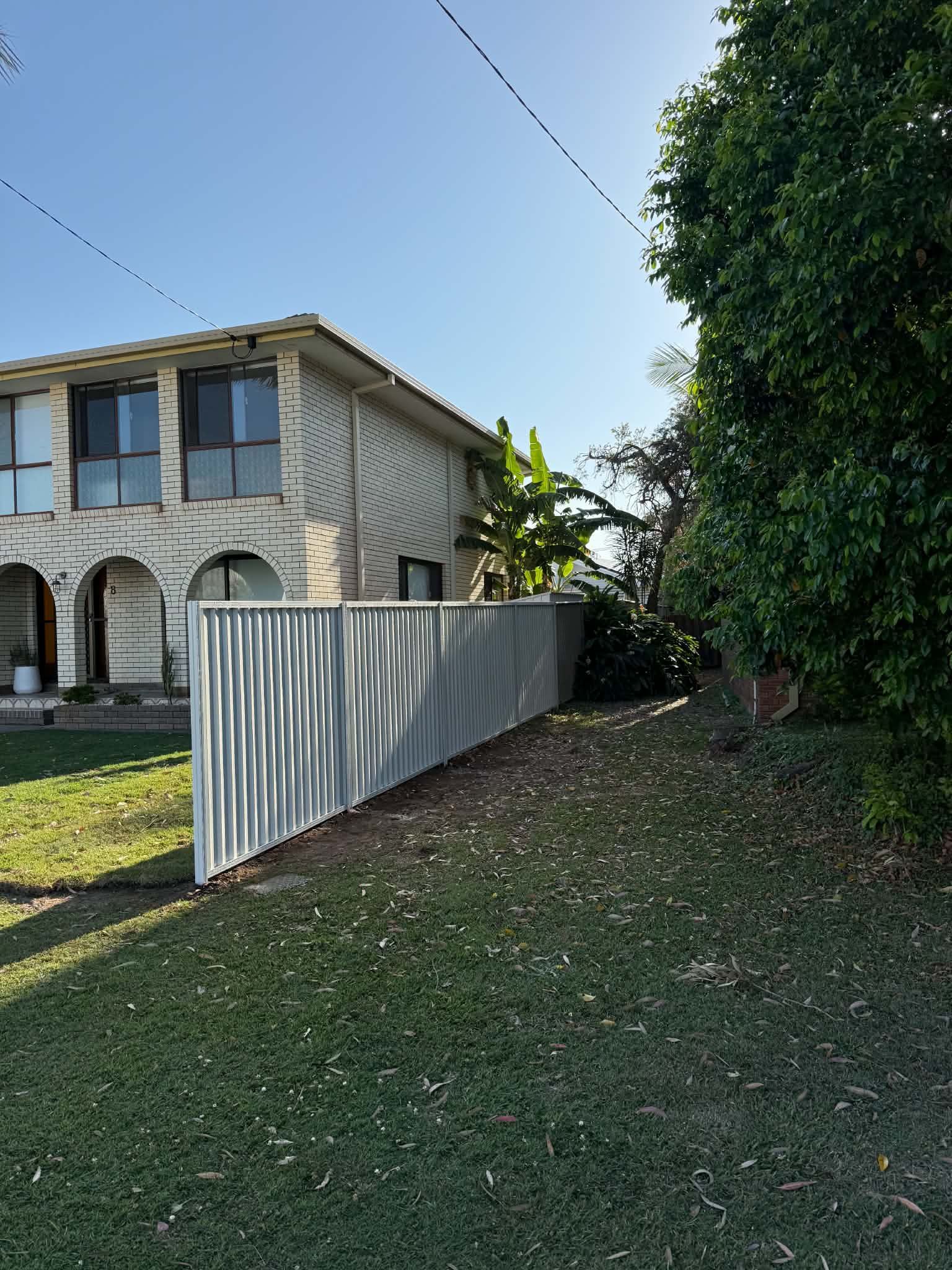 White metal fence beside a grassy yard with a stone house and large tree under a blue sky