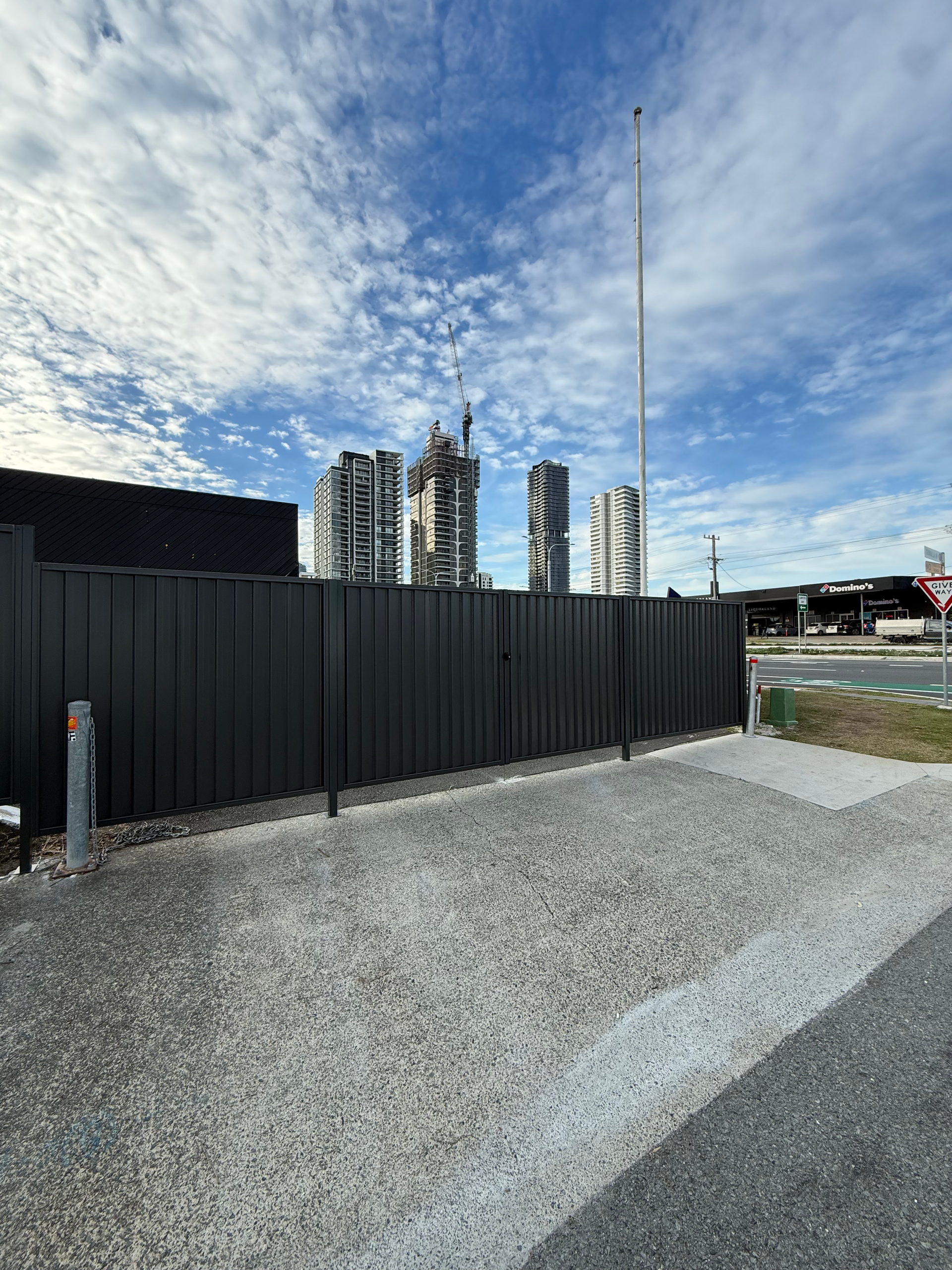 Gravel lot beside a black fence, with tall apartment buildings under a cloudy blue sky.