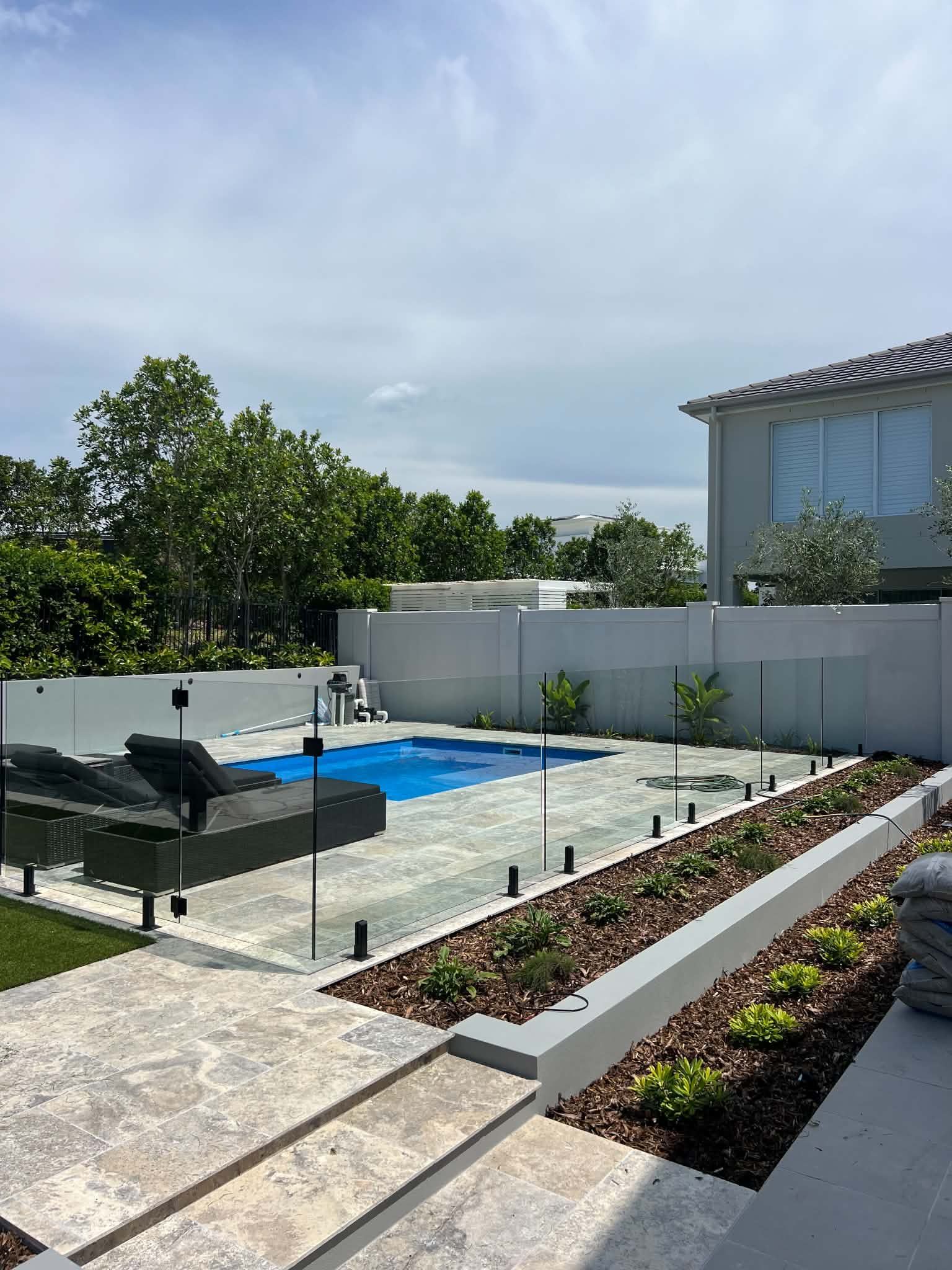 Modern backyard with a blue pool, patio lounge chair, glass fence, and raised planter beds under a cloudy sky