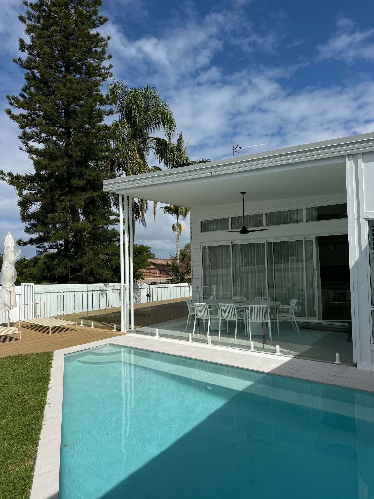 Modern white patio overlooking a turquoise pool, with a covered outdoor dining area and tall trees nearby