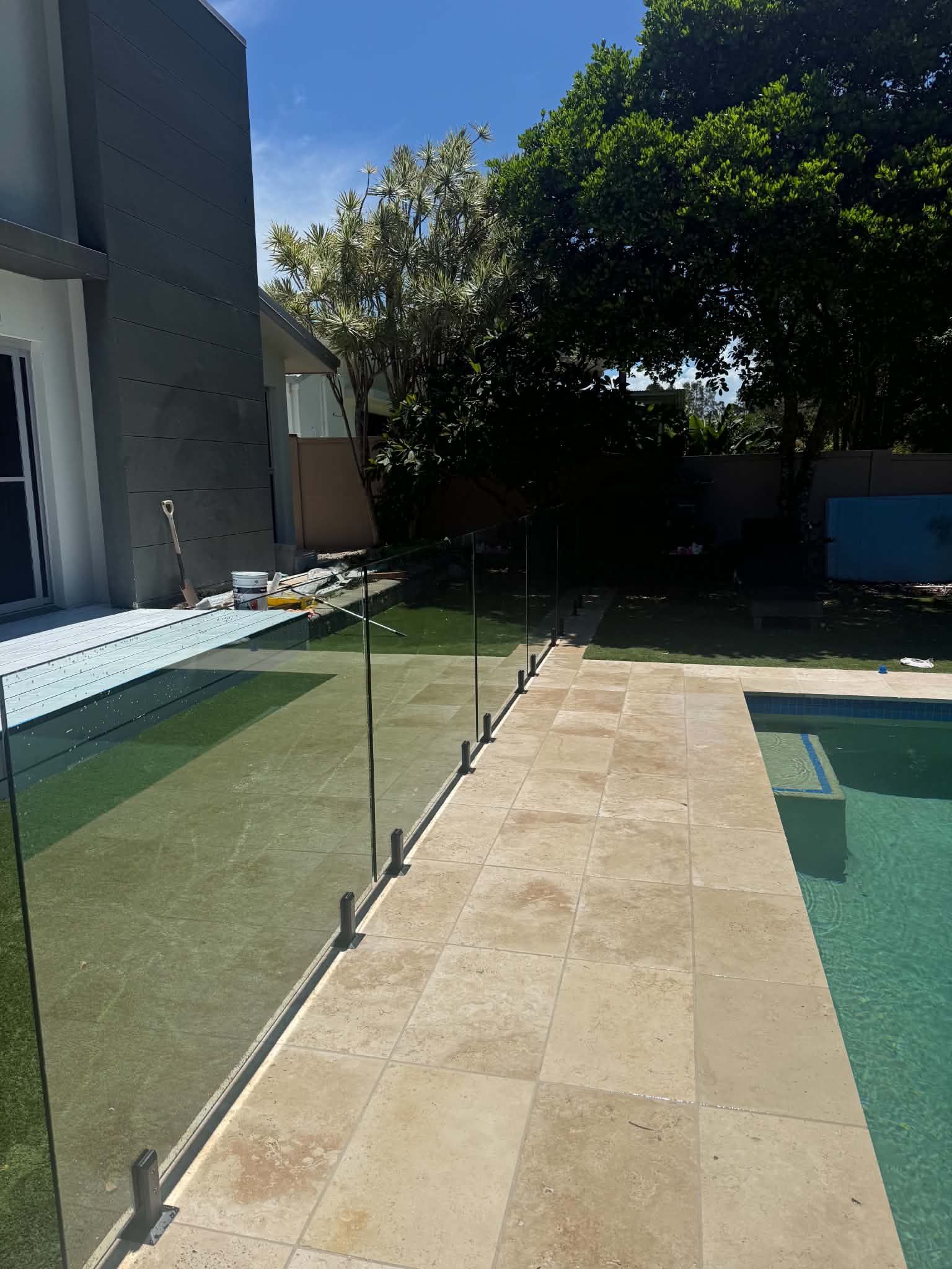 Backyard walkway beside a pool, with green lawn, glass fence, and shaded trees.