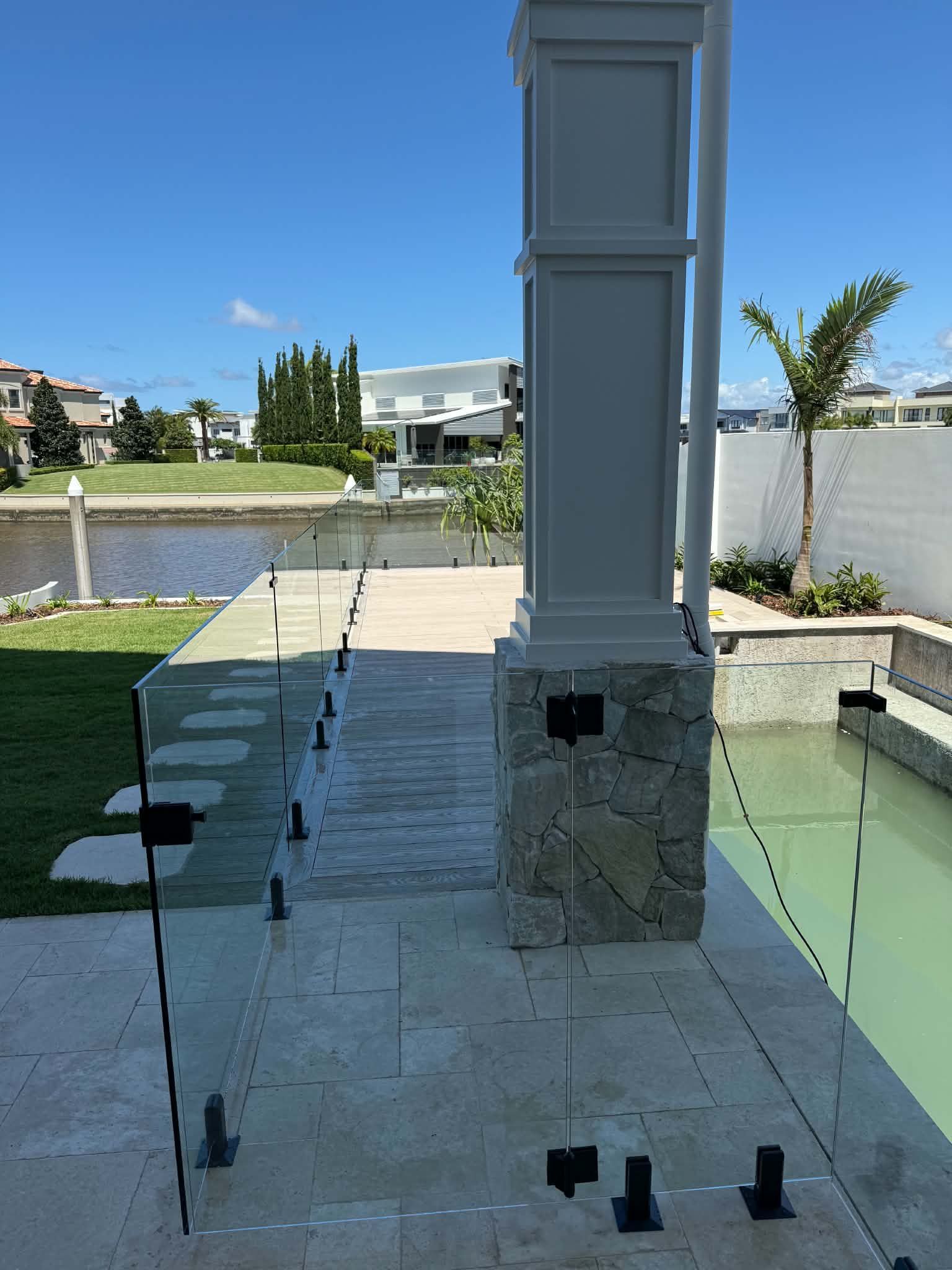 Glass railing beside a pool and waterway, with a white column and stone deck in bright sunlight