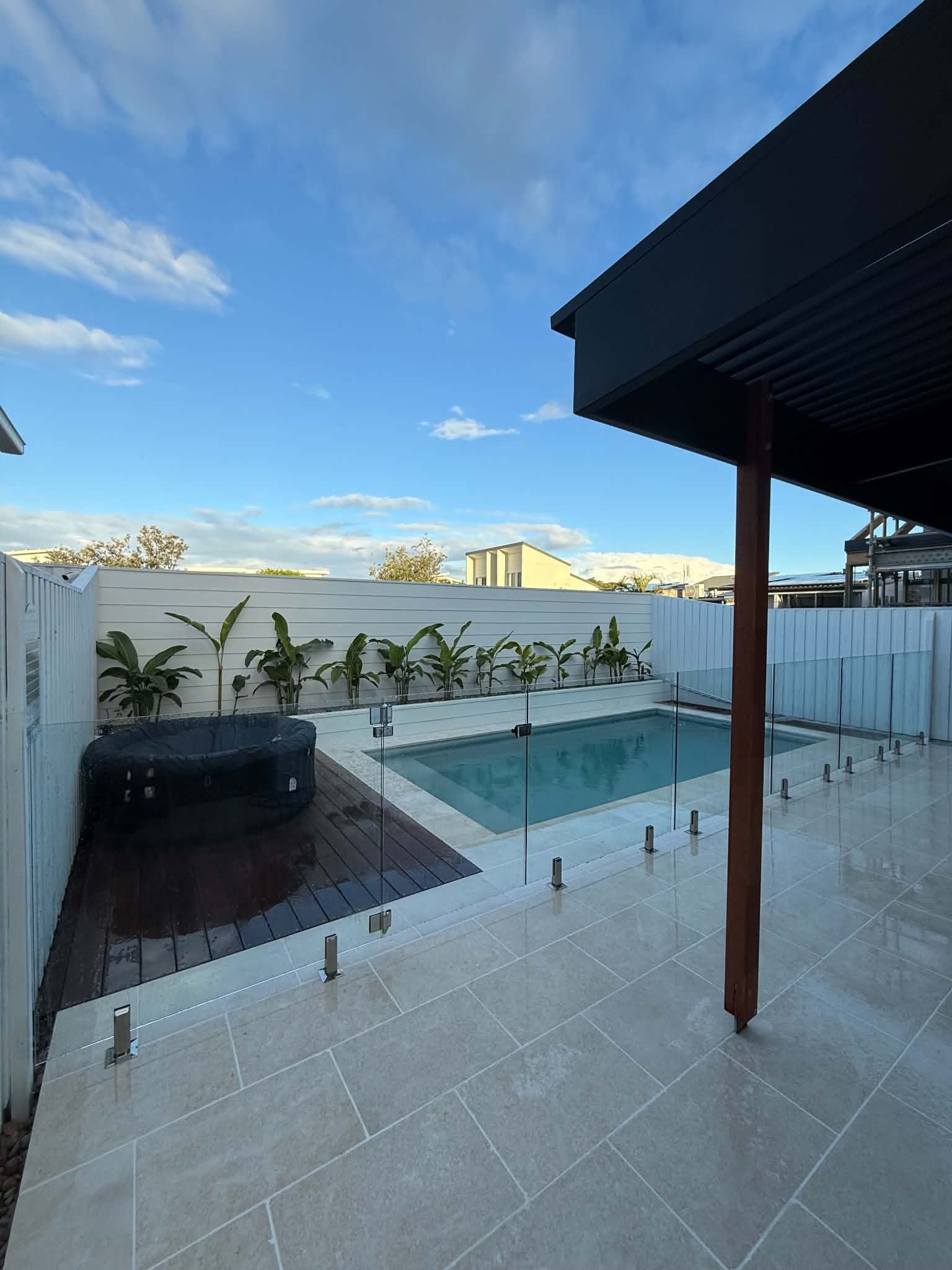 Backyard patio with a small pool, hot tub, white fence, and city skyline under a blue sky