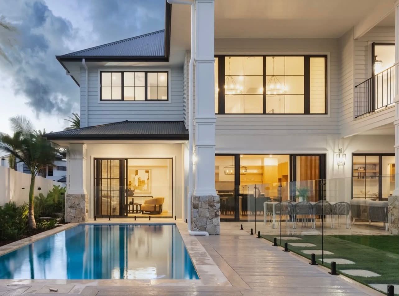 Modern two-story house with lit interior, glass patio doors, and a swimming pool at dusk