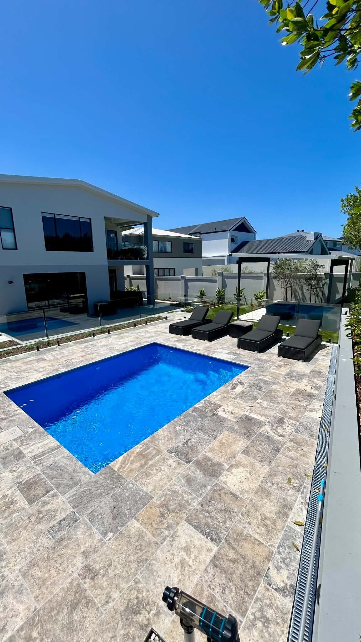 Modern backyard with a blue swimming pool, stone patio, lounge chairs, and white houses under a clear sky