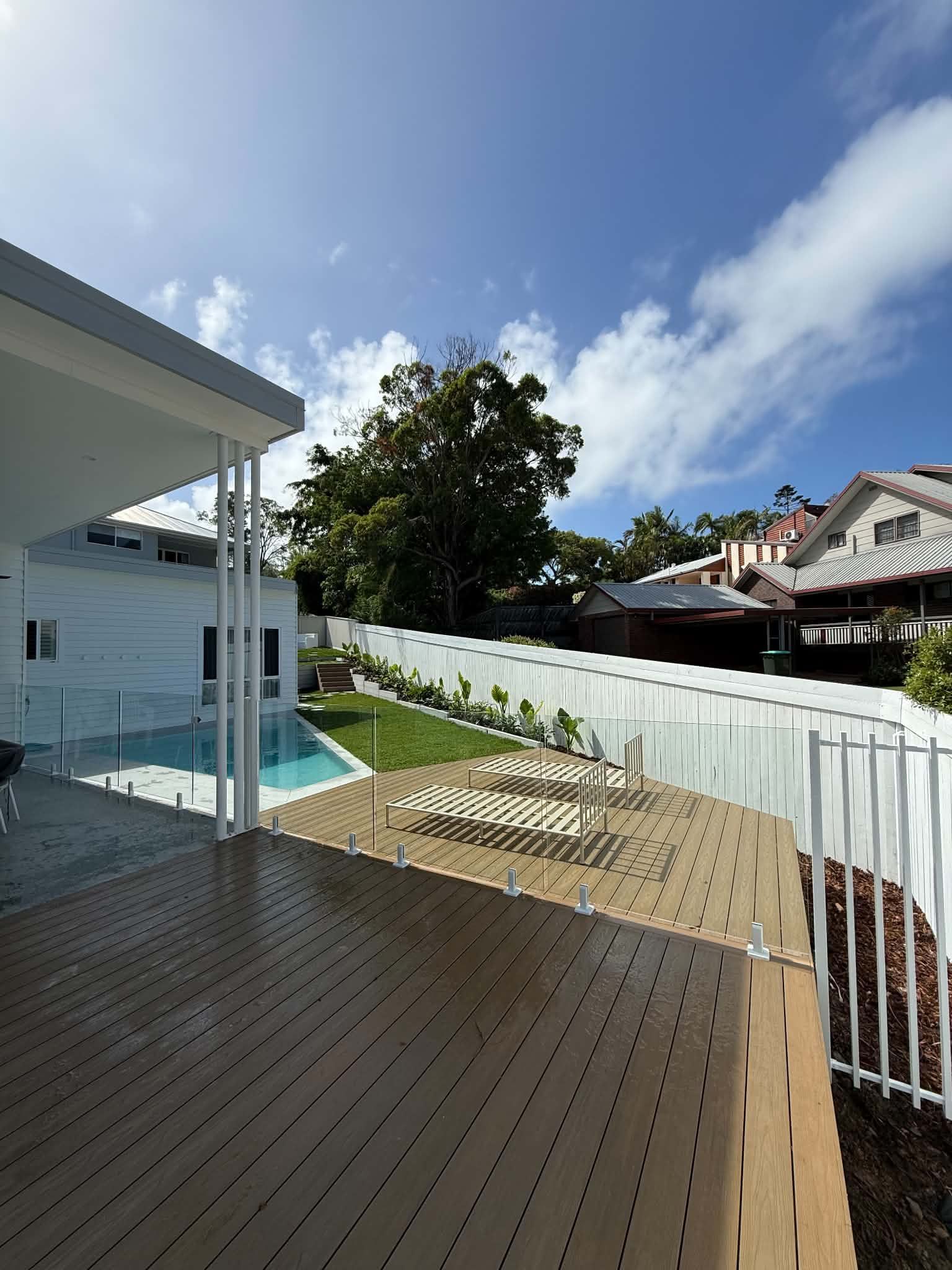 Sunny backyard deck with wooden flooring, white fence, trees, and neighboring houses under a blue sky.