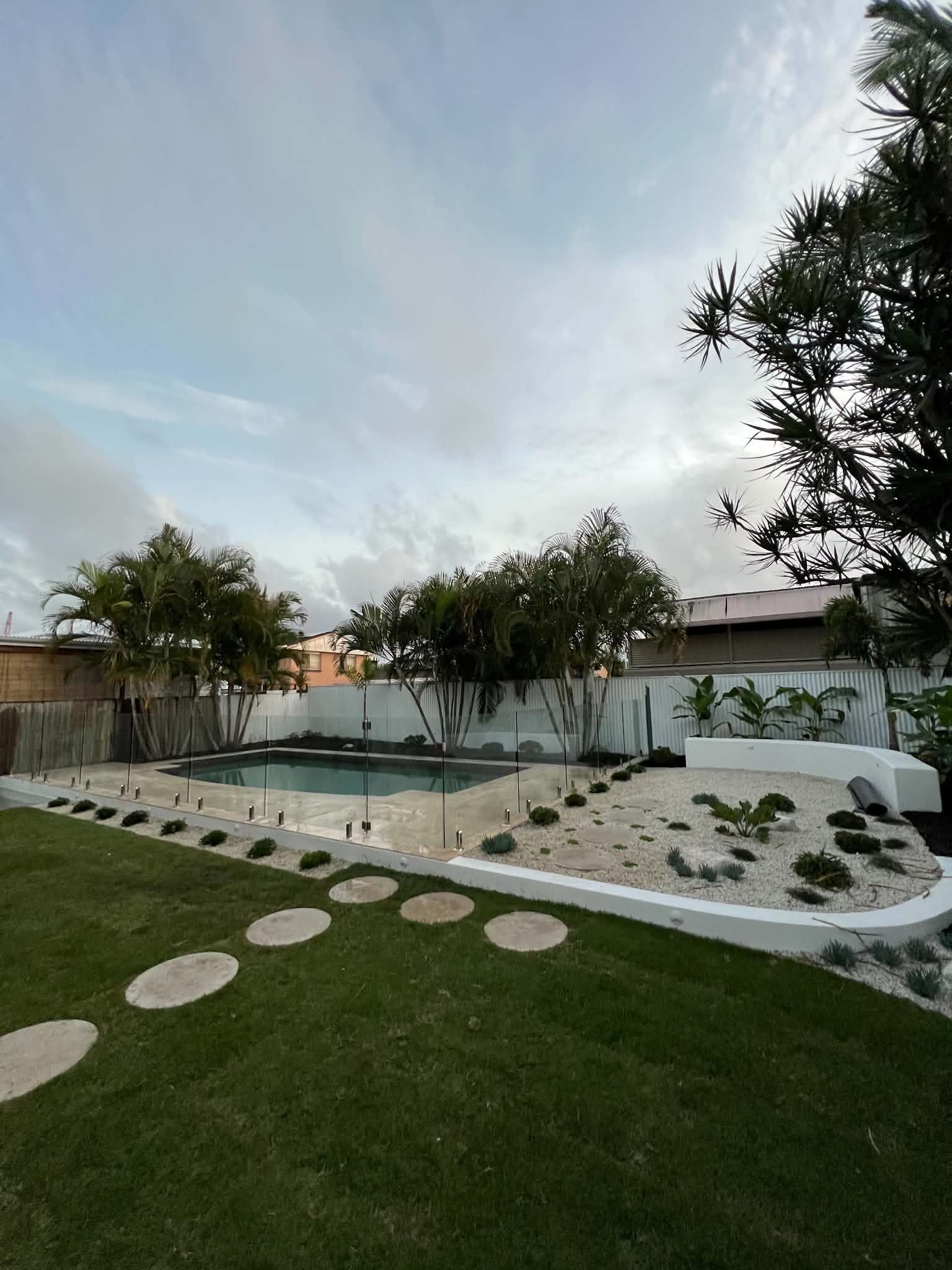 Modern backyard with lawn, stepping stones, white gravel, and trees beside a white wall at dusk