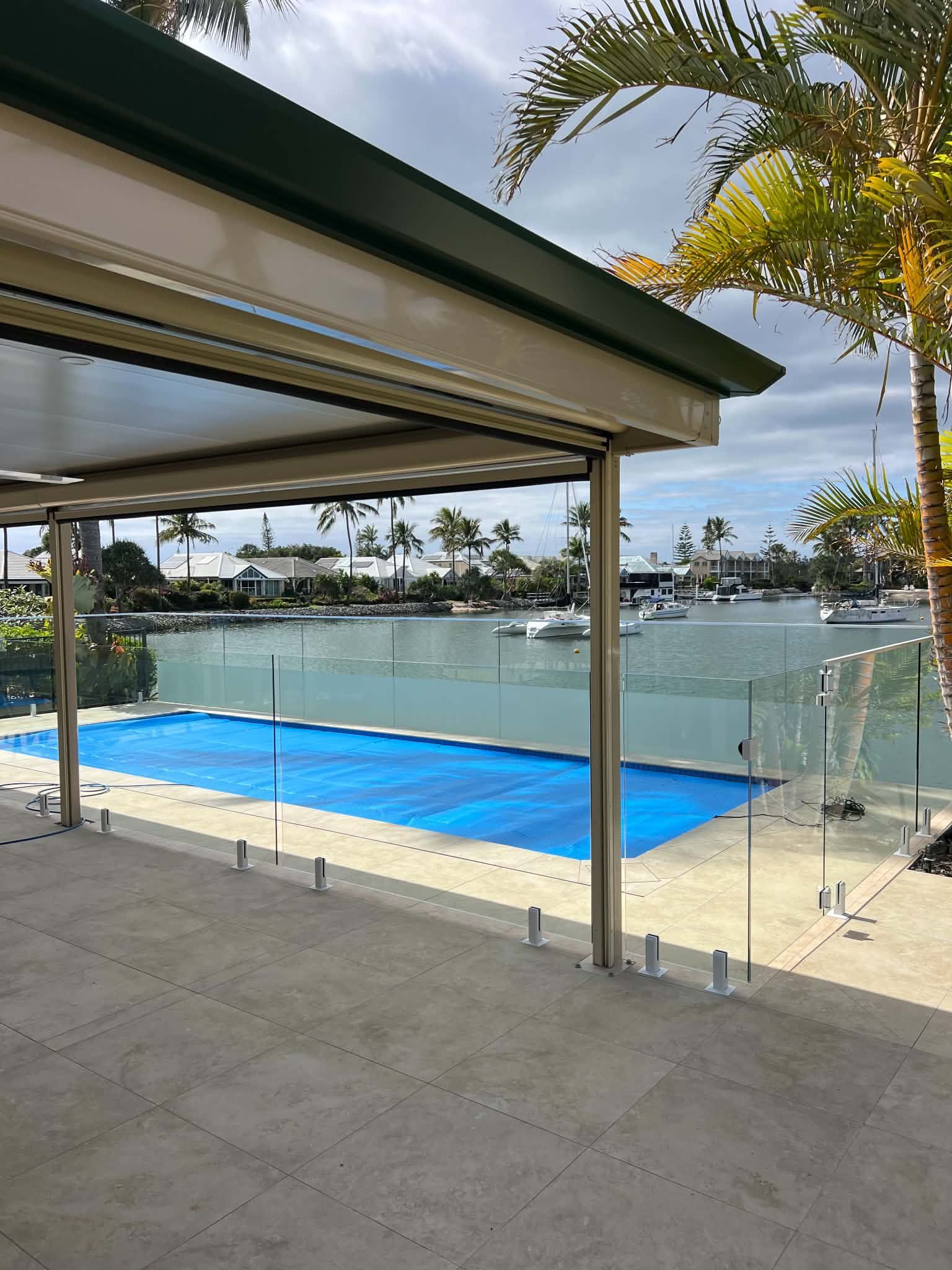 Covered patio overlooking a blue pool and marina, with glass railing and palm trees beyond.