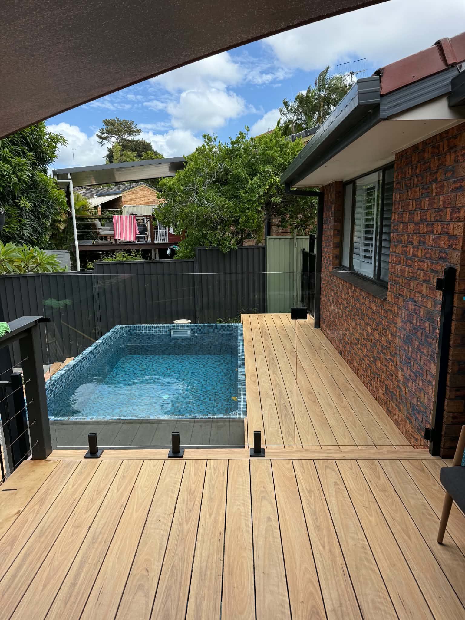 Wooden deck beside a small backyard pool next to a brick house, with a fence and trees beyond