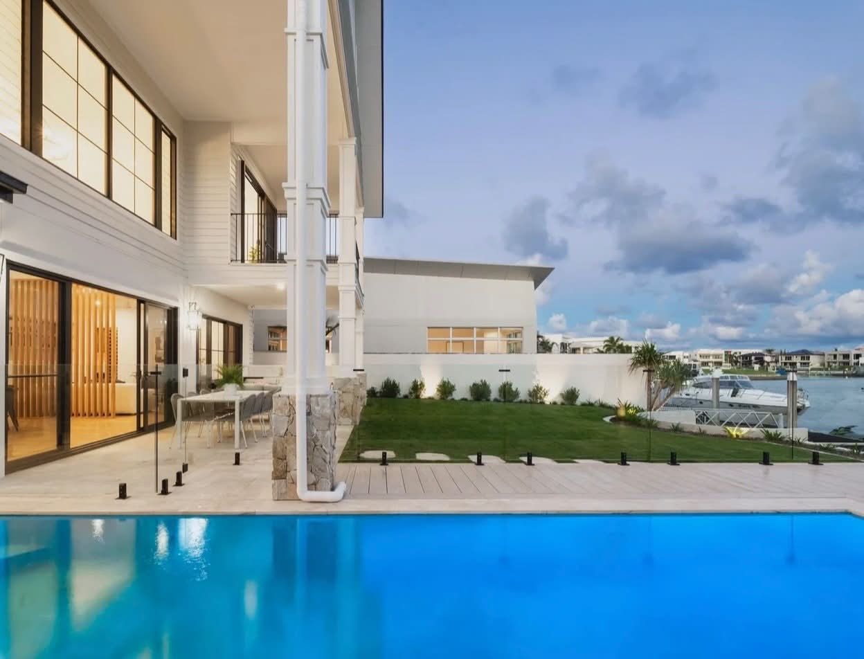 Modern waterfront villa with lit patio, green lawn, and blue infinity pool at dusk